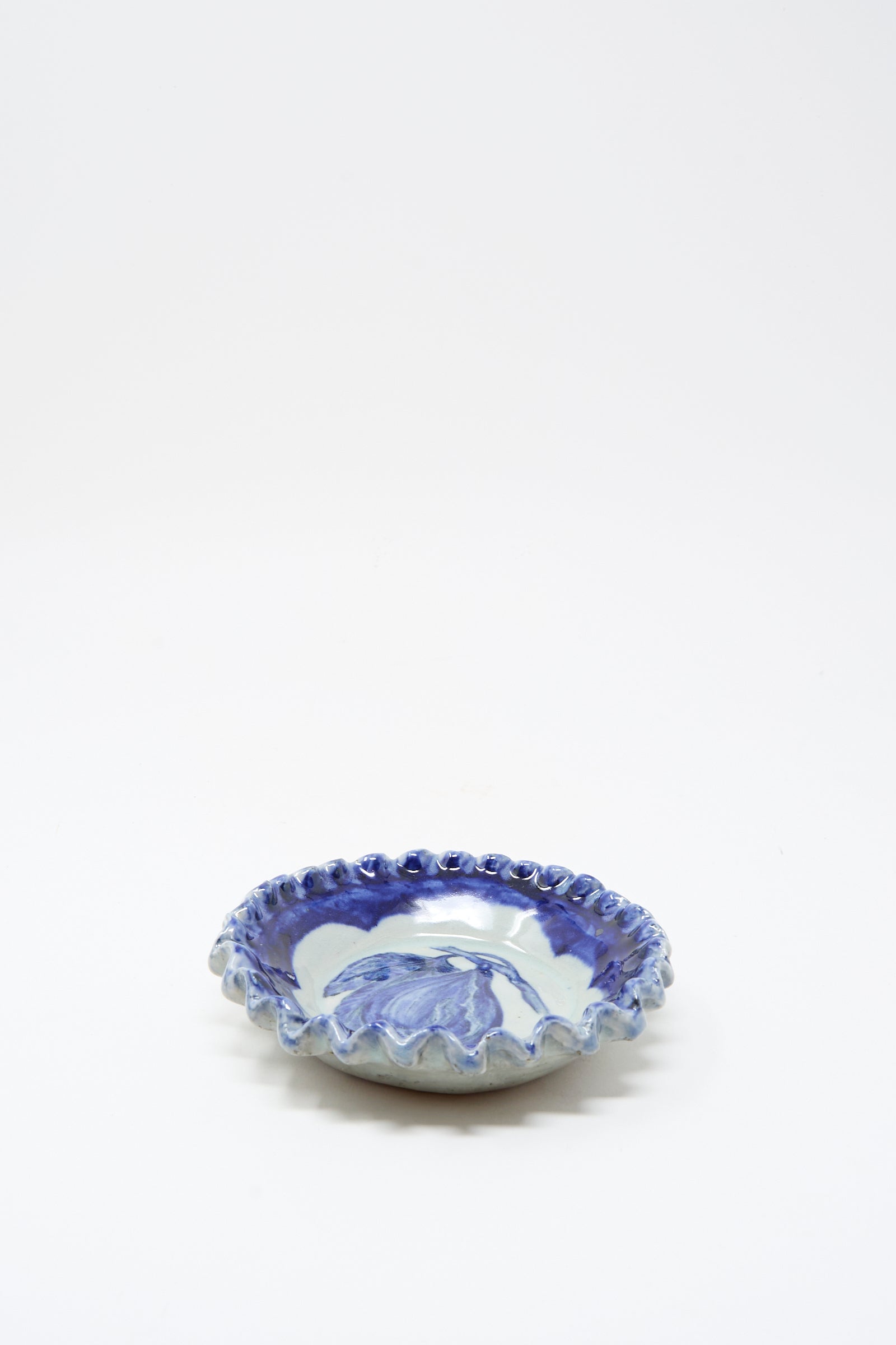 The Ariel Clute Rustic Fig Plate is a shallow, round ceramic dish with a scalloped edge, hand painted with blue floral patterns on a white porcelain background.