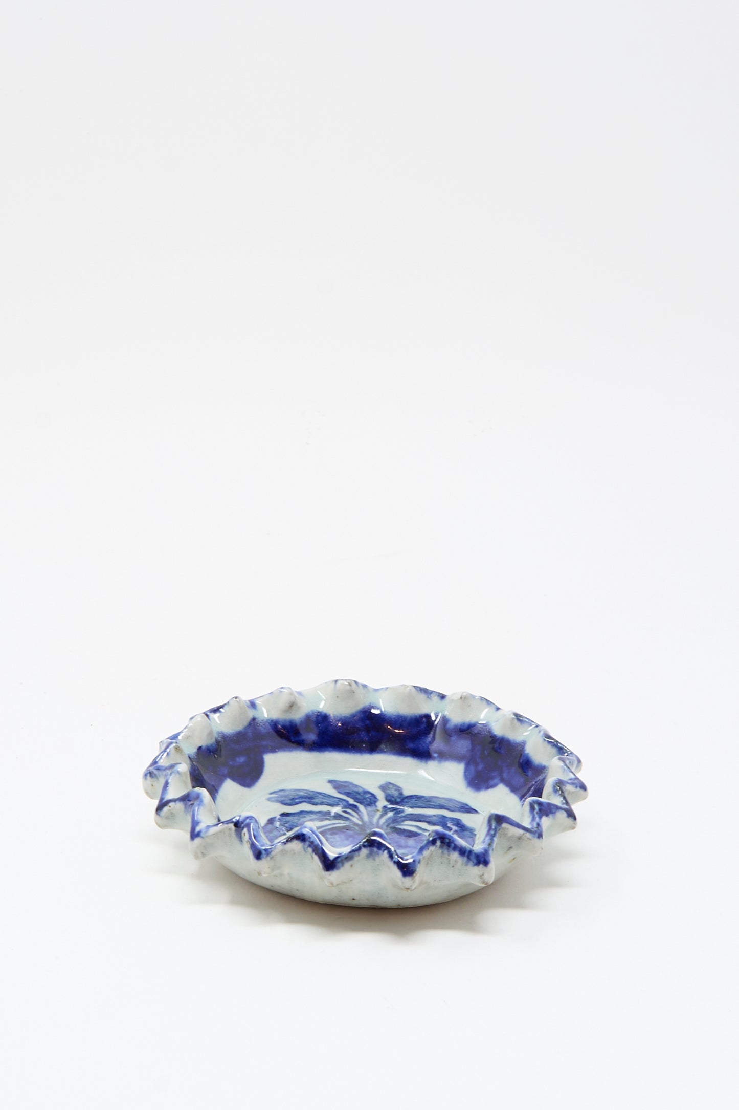 The Rustic Turnip Bowl by Ariel Clute is a handmade shallow ceramic dish with a ruffled edge and blue floral patterns on white, shown centered against a plain white surface.