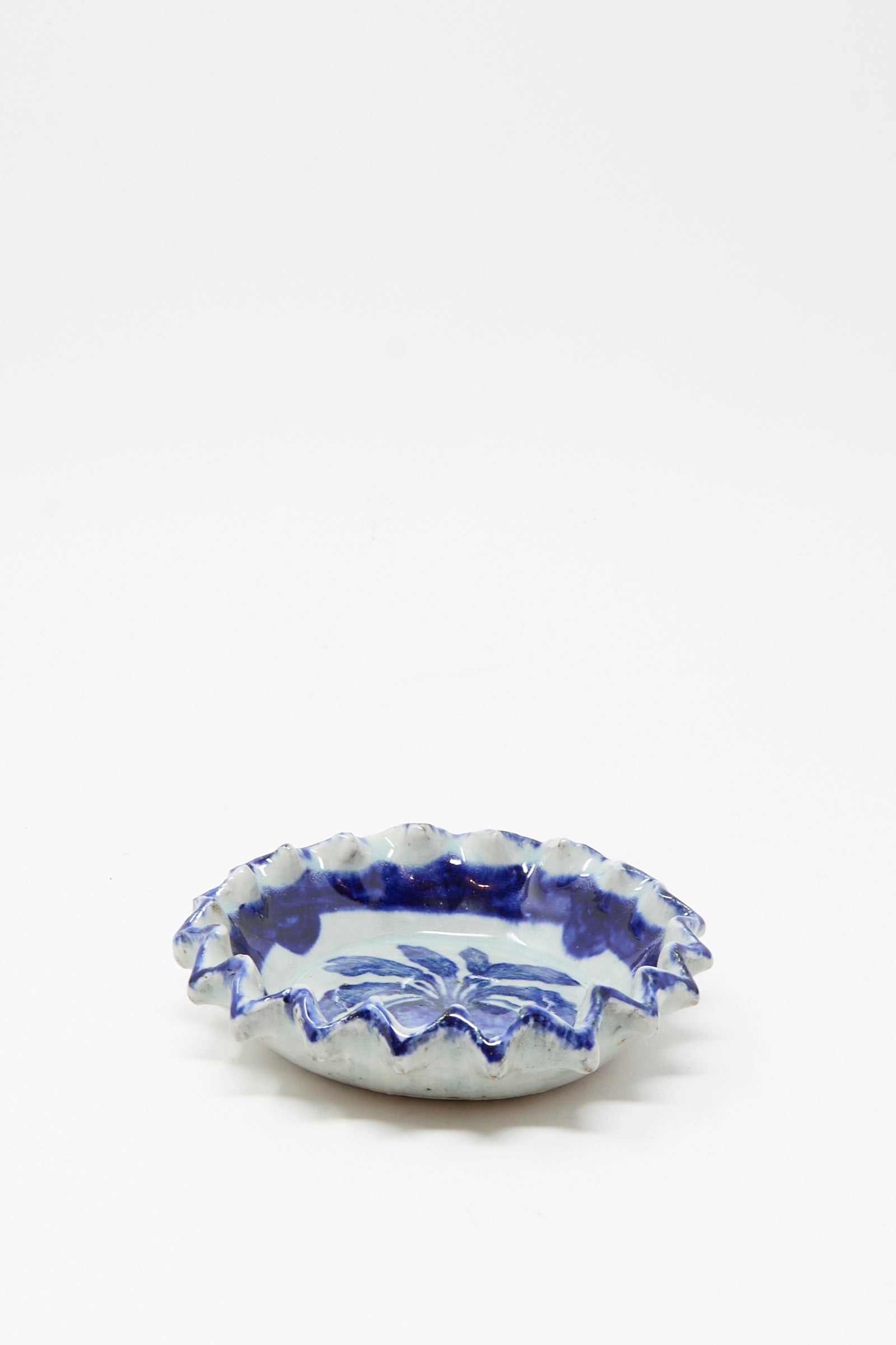 The Rustic Turnip Bowl by Ariel Clute is a handmade shallow ceramic dish with a ruffled edge and blue floral patterns on white, shown centered against a plain white surface.