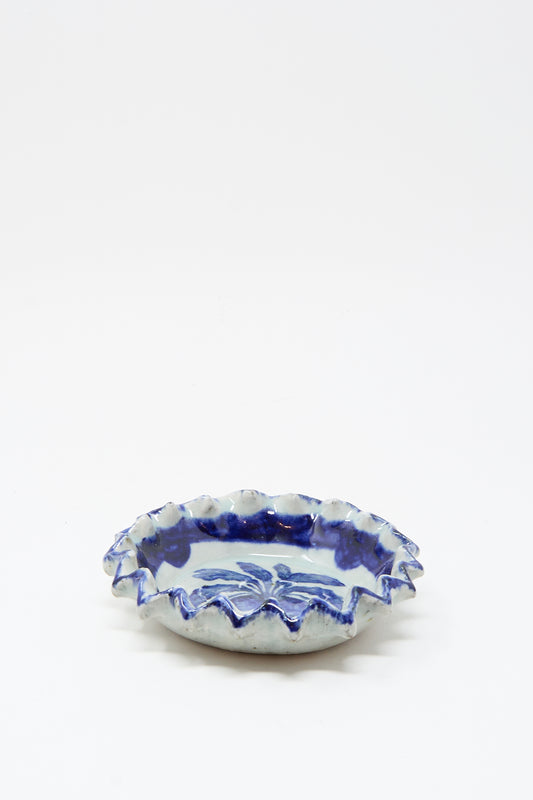 The Rustic Turnip Bowl by Ariel Clute is a handmade shallow ceramic dish with a ruffled edge and blue floral patterns on white, shown centered against a plain white surface.