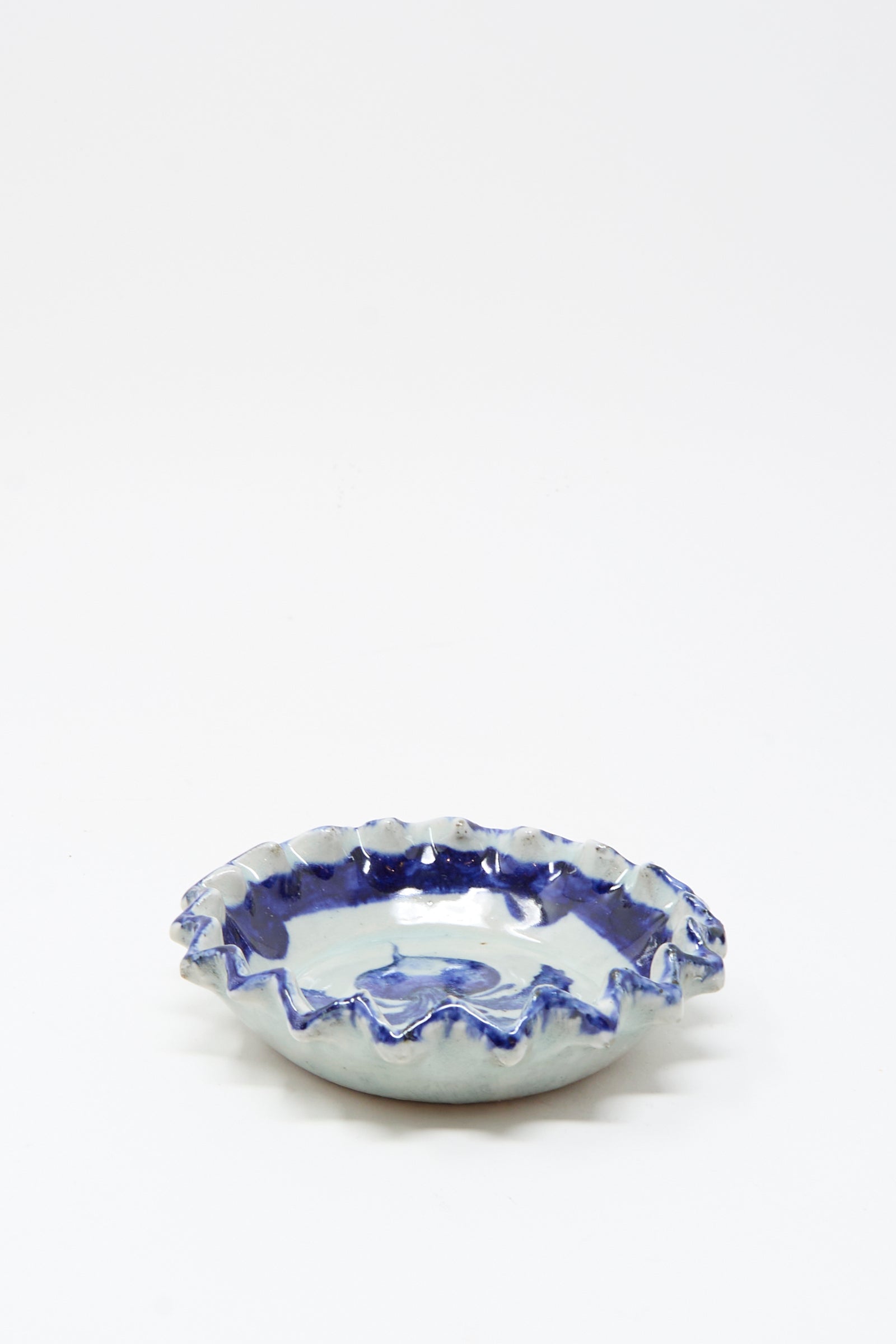 The Rustic Turnip Bowl by Ariel Clute is a shallow ceramic dish with a wavy ruffled edge and blue-and-white glaze, featuring a hand-painted porcelain plate style, shown on a plain white background.