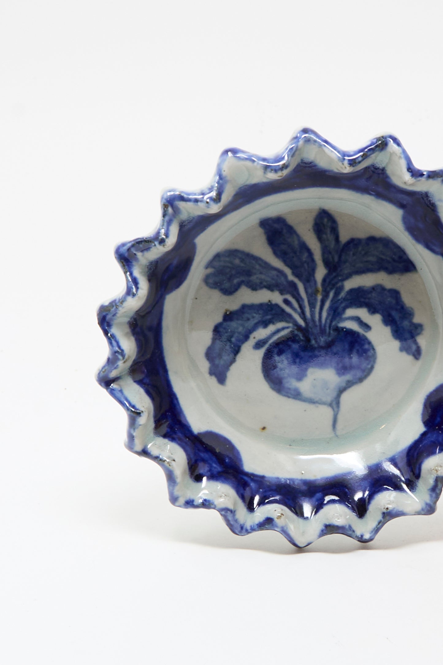 The Rustic Turnip Bowl by Ariel Clute is a ceramic dish with wavy edges, showcasing a hand-painted blue turnip design on a white porcelain center.