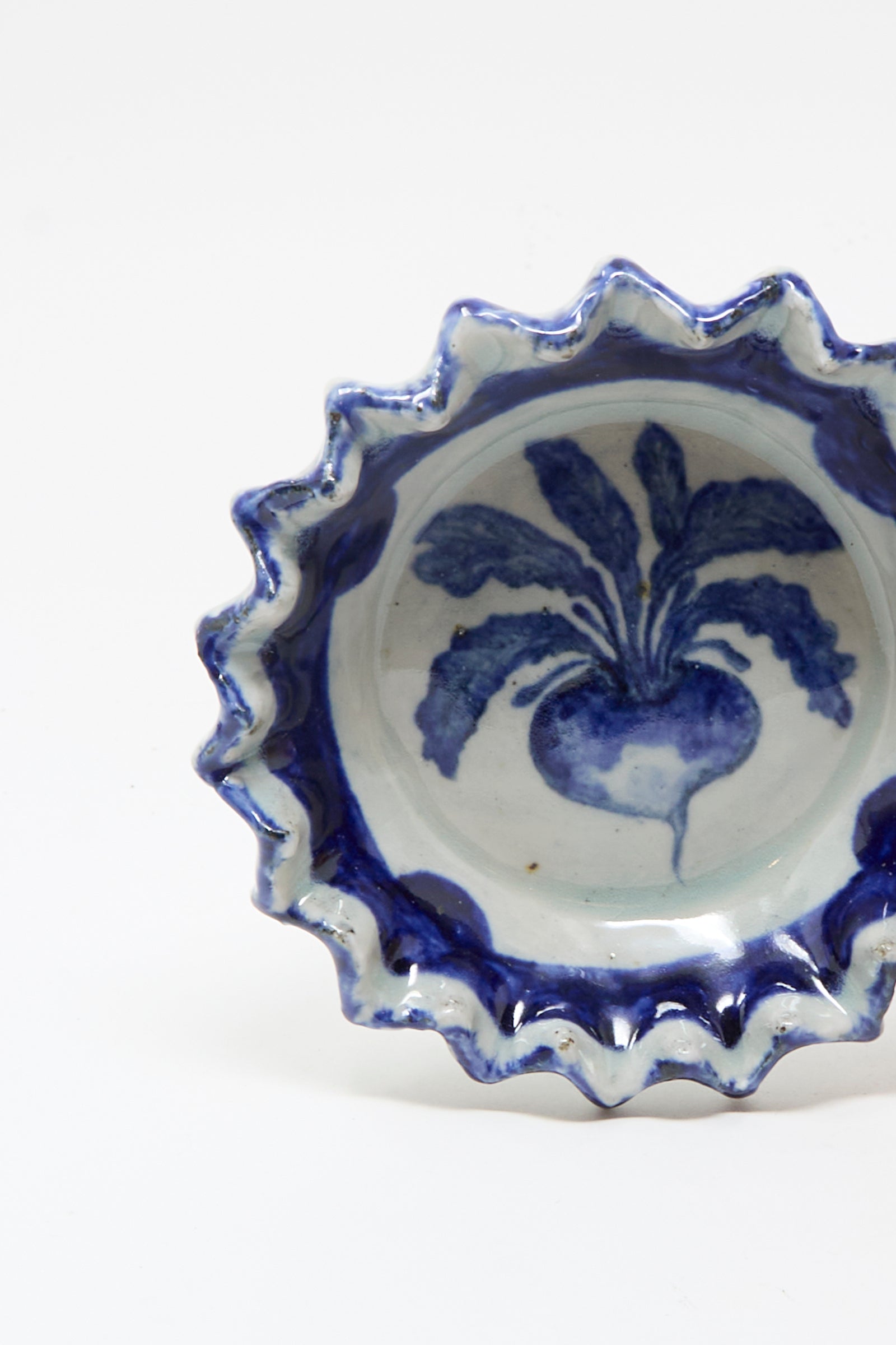 The Rustic Turnip Bowl by Ariel Clute is a ceramic dish with wavy edges, showcasing a hand-painted blue turnip design on a white porcelain center.