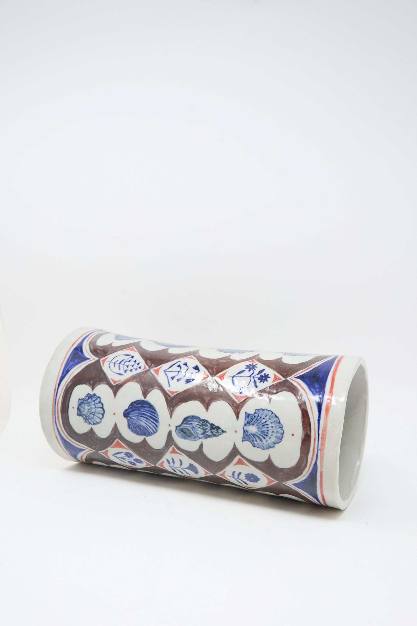 The Sea Shore Vessel by Ariel Clute is a cylindrical ceramic vase featuring blue, brown, and red floral and geometric patterns, shown lying on its side against a plain white background.
