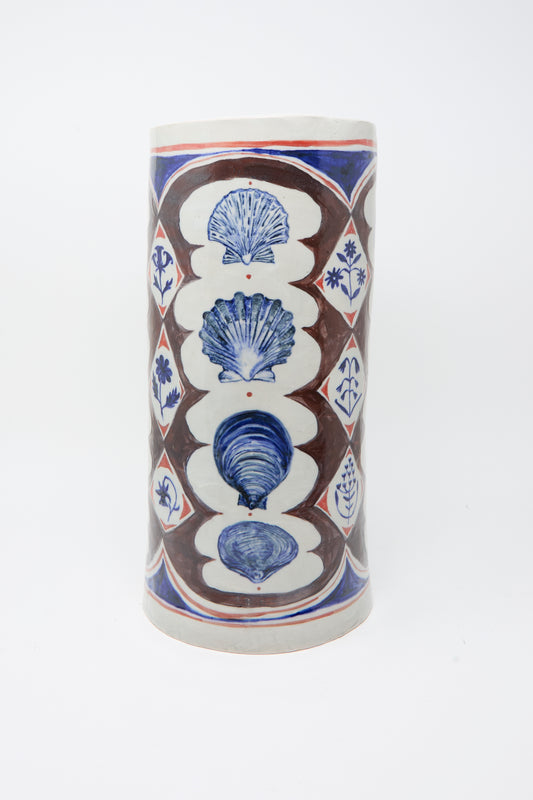 The Sea Shore Vessel by Ariel Clute is a tall, hand-painted porcelain piece with shell motifs and geometric floral patterns in brown, blue, and red on a white background.