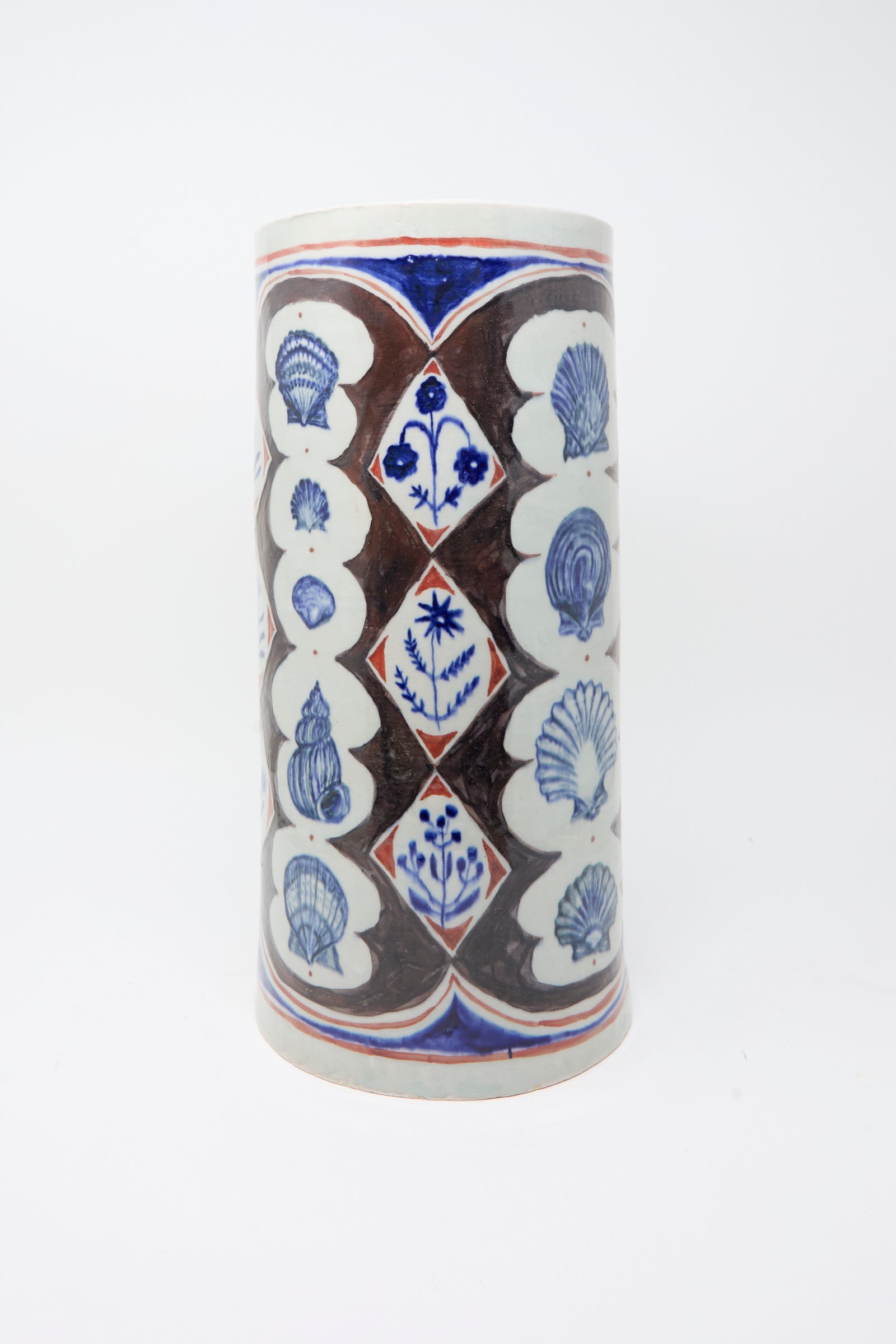 The Sea Shore Vessel by Ariel Clute is a cylindrical, hand-painted porcelain piece featuring blue floral and shell designs set in geometric panels on a white background.