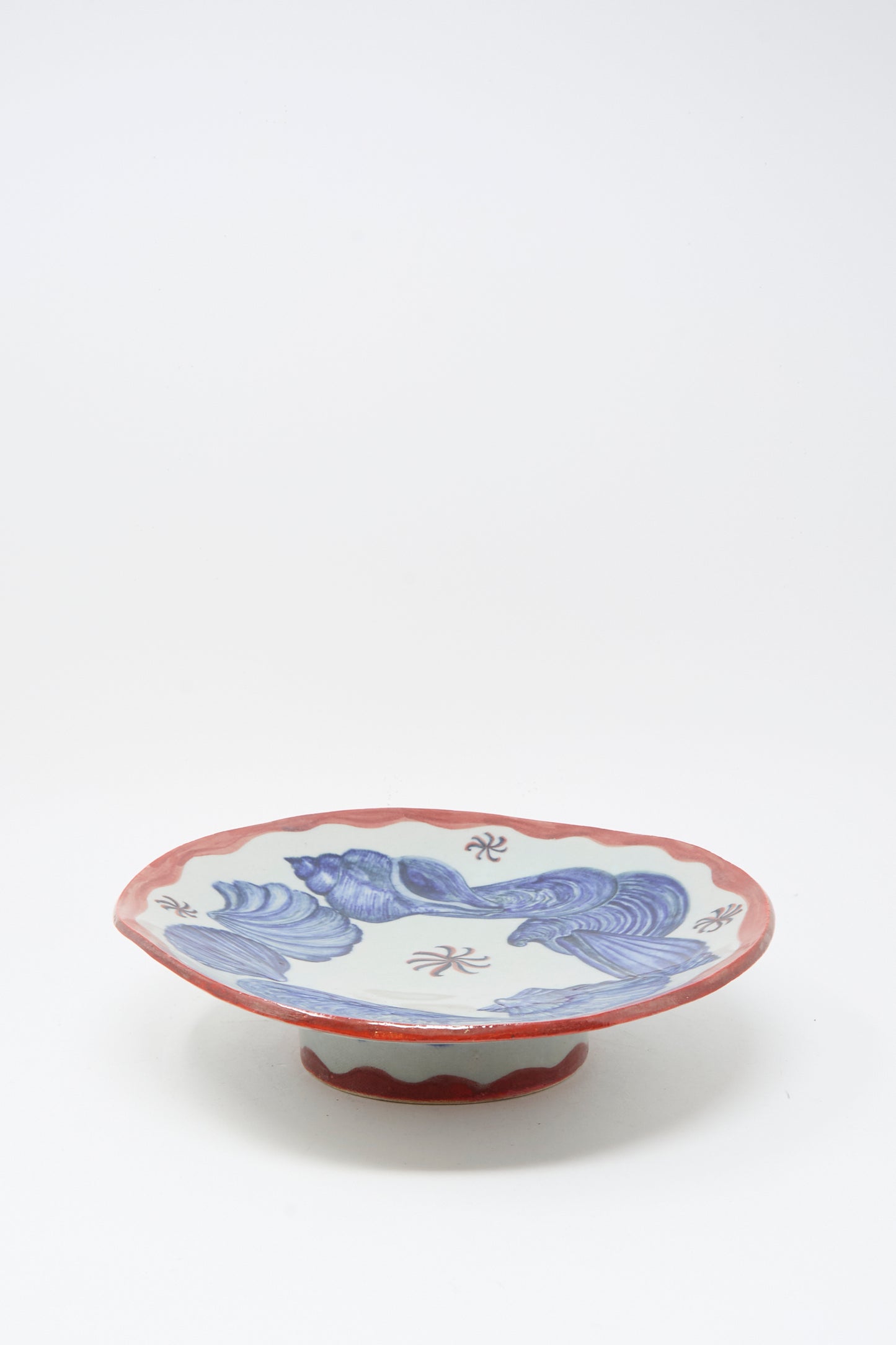 The Shell Pedestal by Ariel Clute is a hand-painted porcelain plate featuring a red scalloped rim, blue wave motifs, and Chinese characters on a short pedestal base, shown against a white background.