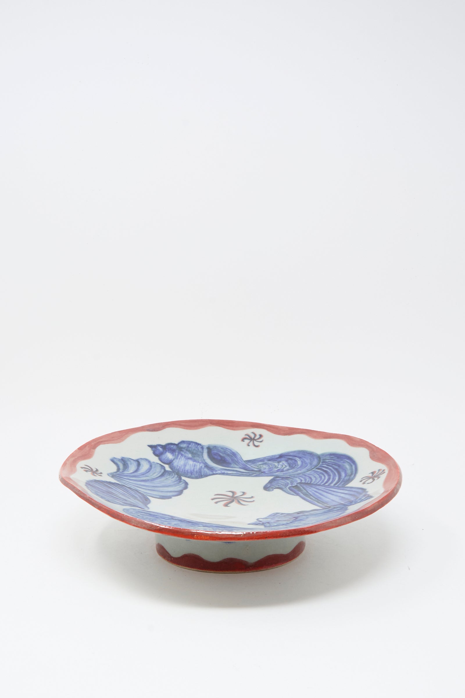 The Shell Pedestal by Ariel Clute is a hand-painted porcelain plate featuring a red scalloped rim, blue wave motifs, and Chinese characters on a short pedestal base, shown against a white background.