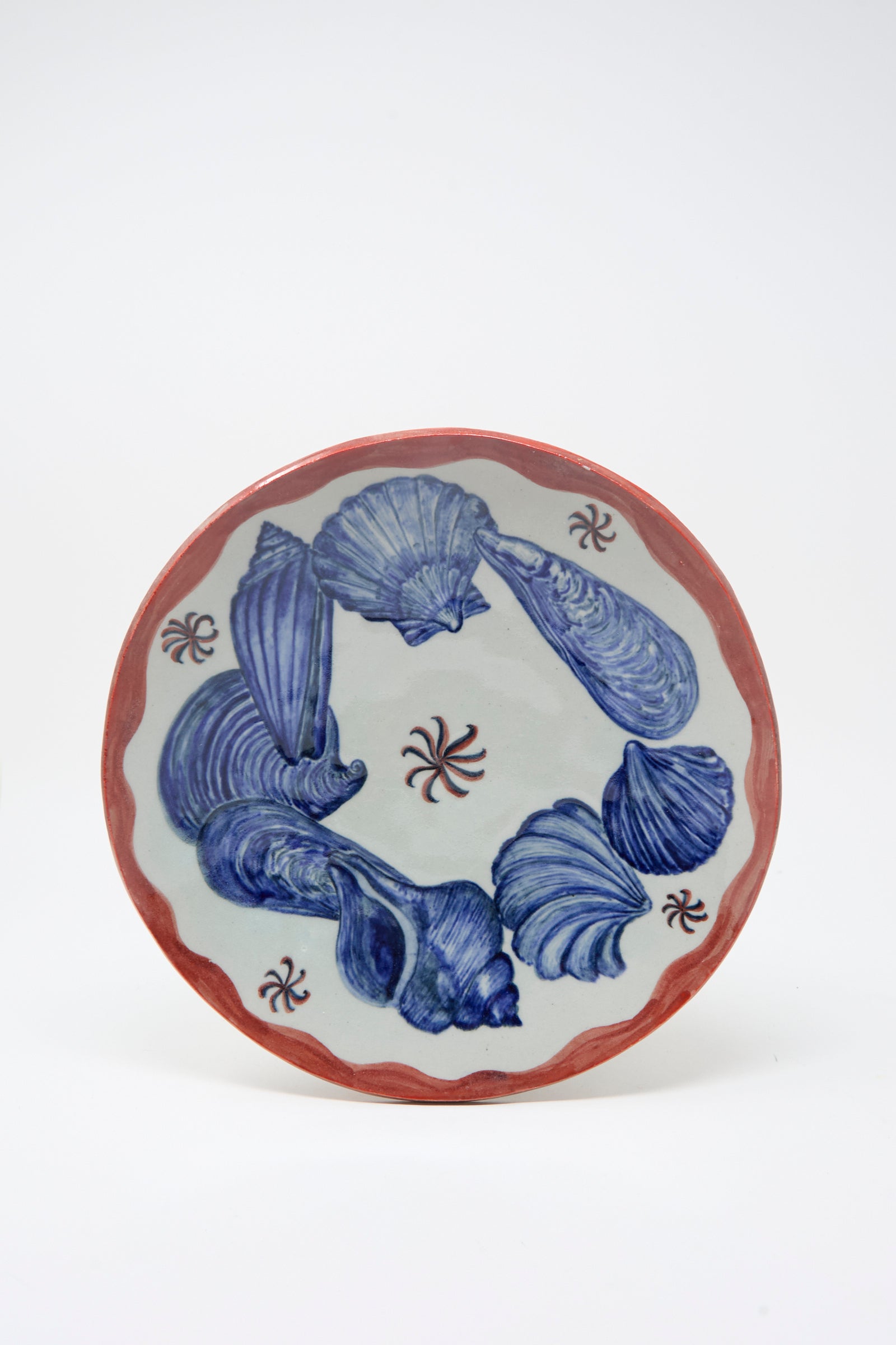 The Shell Pedestal by Ariel Clute is handmade in California with a red rim, blue seashell motifs, and small red spiral patterns, all hand painted on white porcelain.