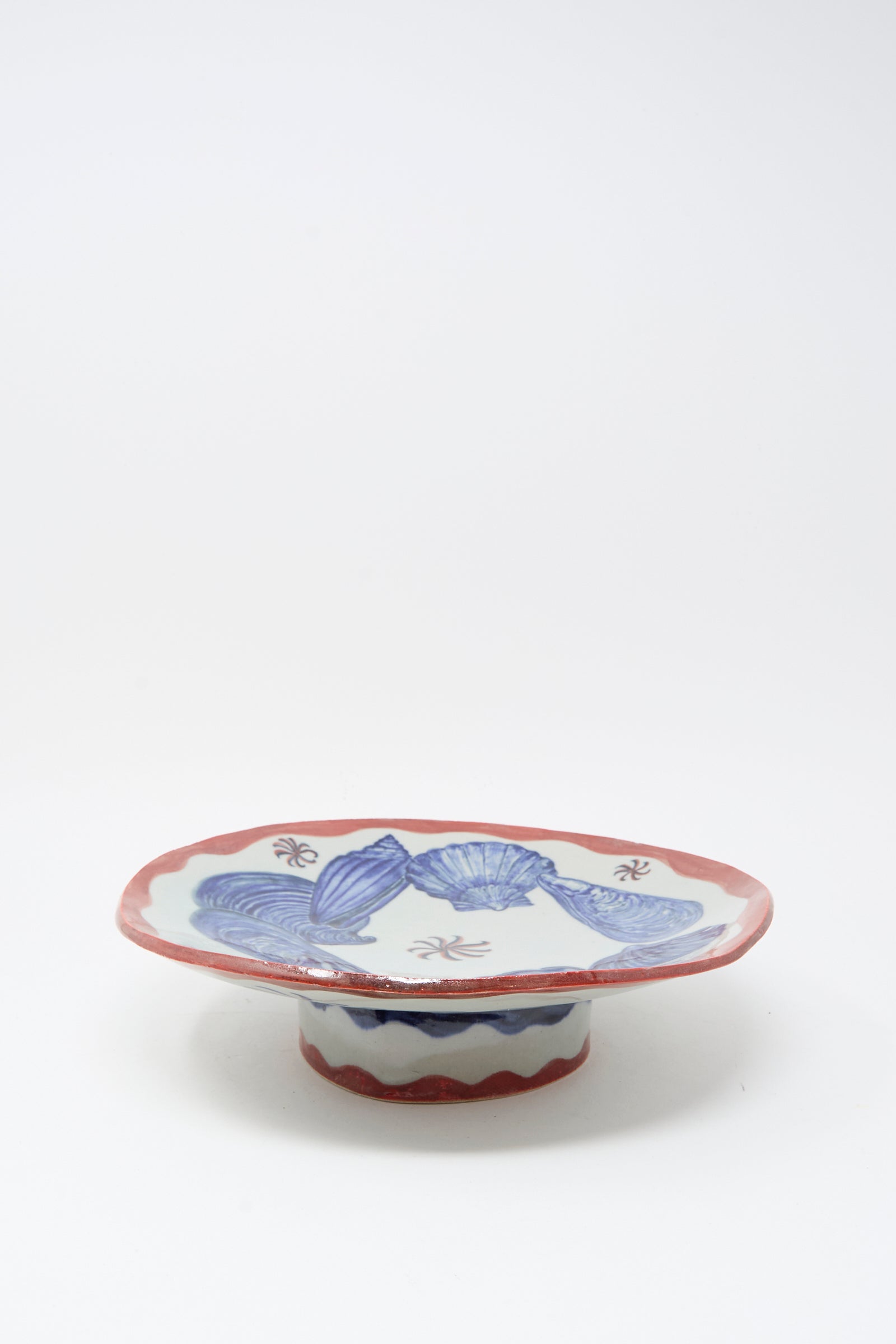 The Shell Pedestal by Ariel Clute is a handmade shallow porcelain plate with a red rim, blue leaf-like designs, and a short base—crafted in California.