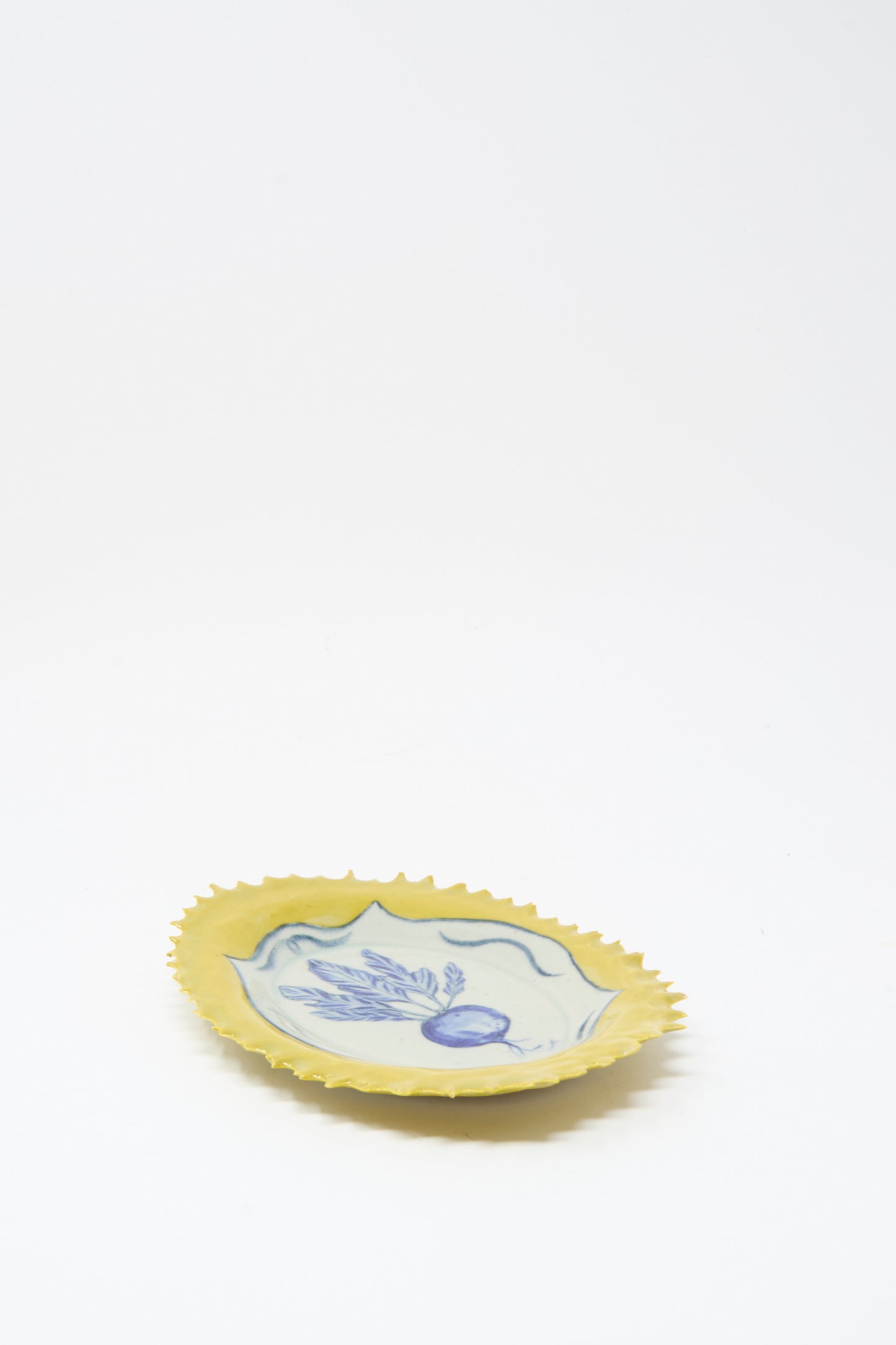 The Ariel Clute Yellow Radish Plate is a handmade yellow ceramic plate with a scalloped edge and blue floral radish design at the center, crafted in California and shown on a plain white background.