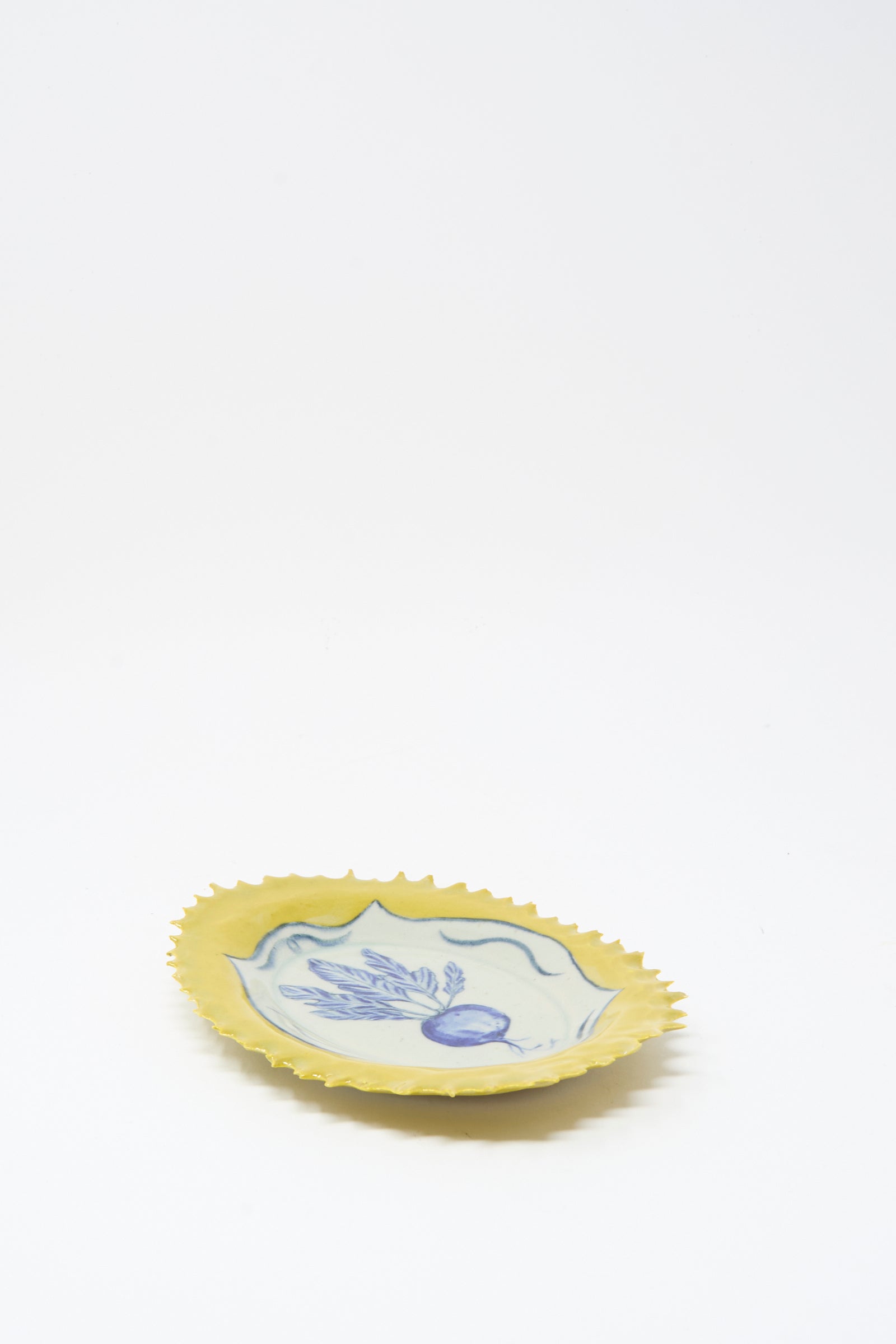 The Ariel Clute Yellow Radish Plate is a handmade yellow ceramic plate with a scalloped edge and blue floral radish design at the center, crafted in California and shown on a plain white background.