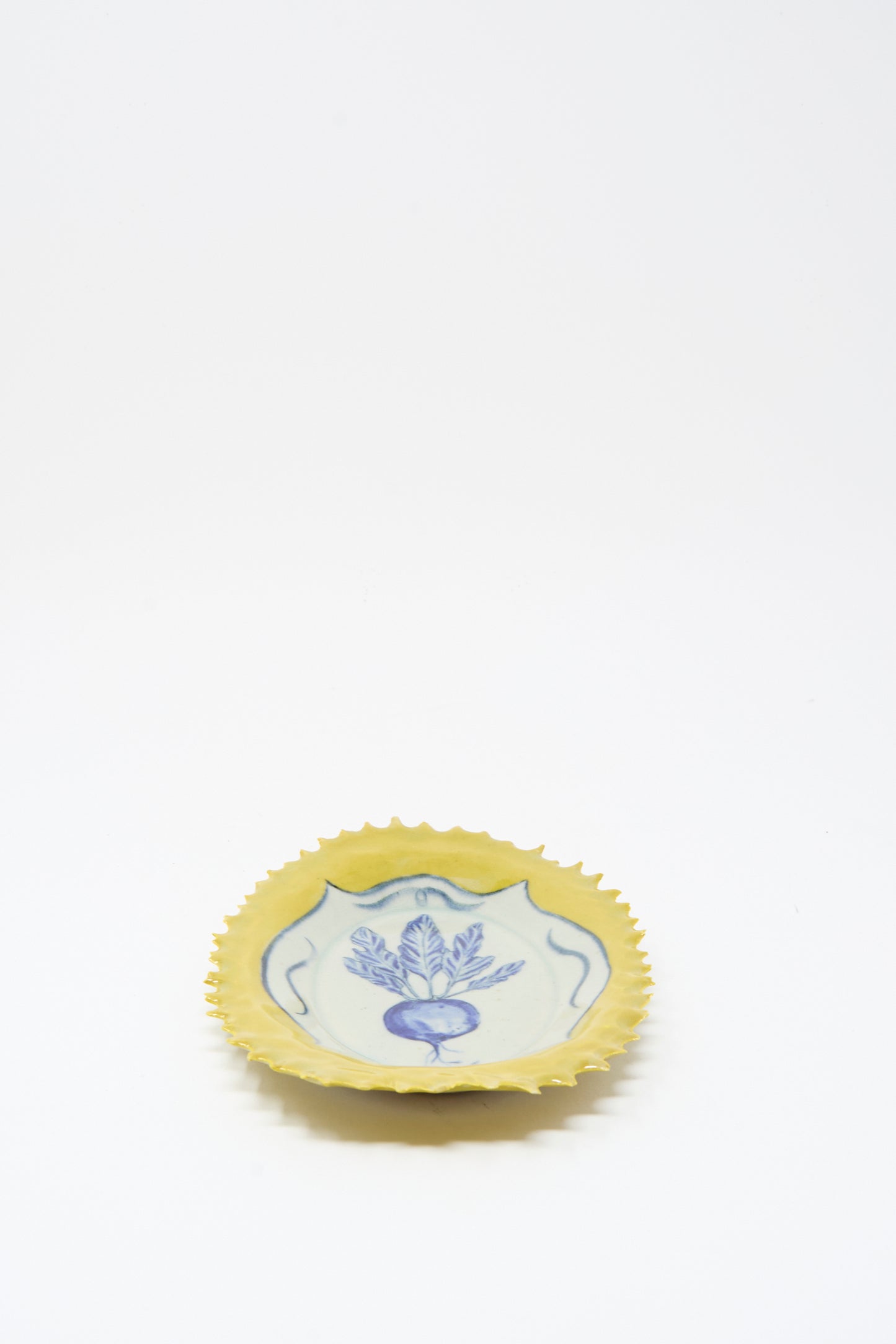 The Yellow Radish Plate by Ariel Clute is a small, round ceramic dish handmade in California, with a yellow scalloped edge and blue floral design on a white background.