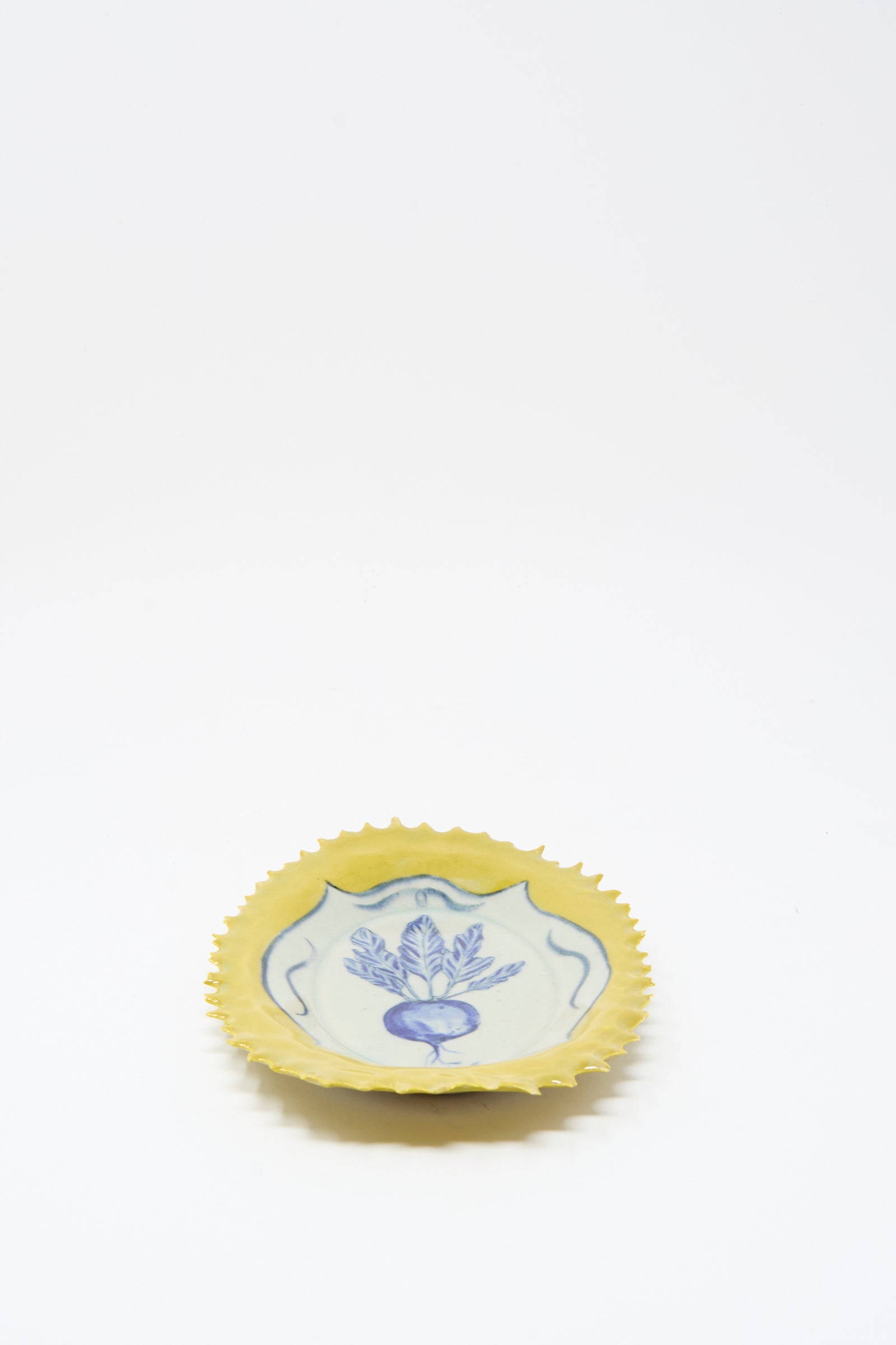 The Yellow Radish Plate by Ariel Clute is a small, round ceramic dish handmade in California, with a yellow scalloped edge and blue floral design on a white background.