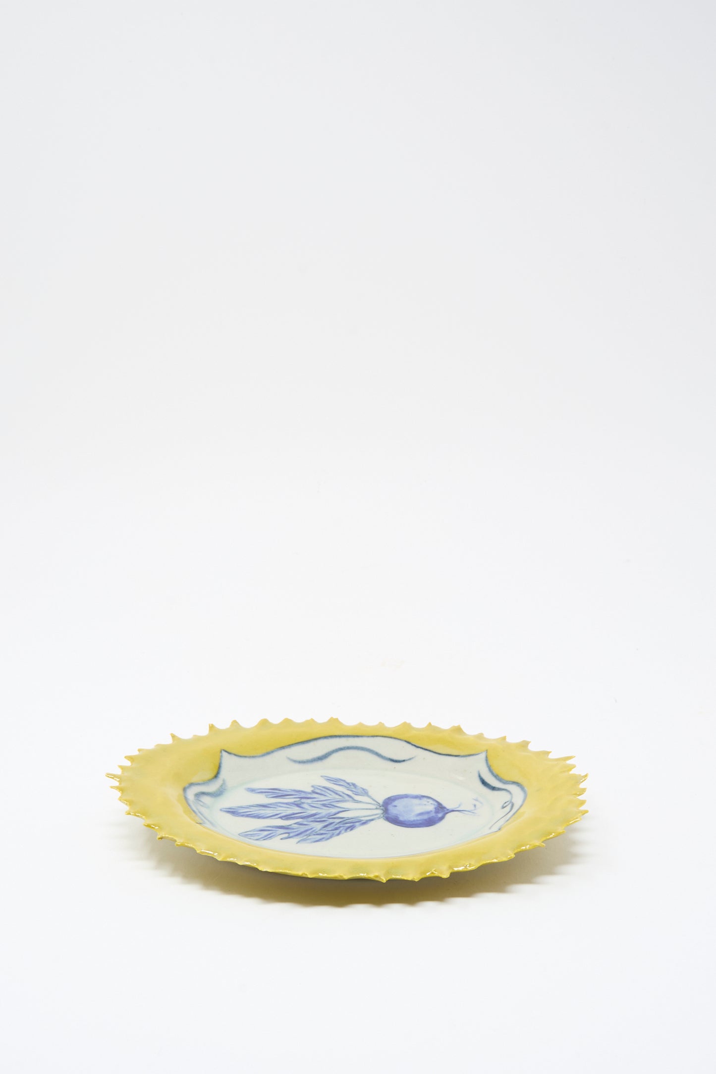 The Yellow Radish Plate by Ariel Clute is a handmade yellow ceramic plate with jagged edges and a blue and white fish illustration at the center, crafted in California, shown on a plain white background.