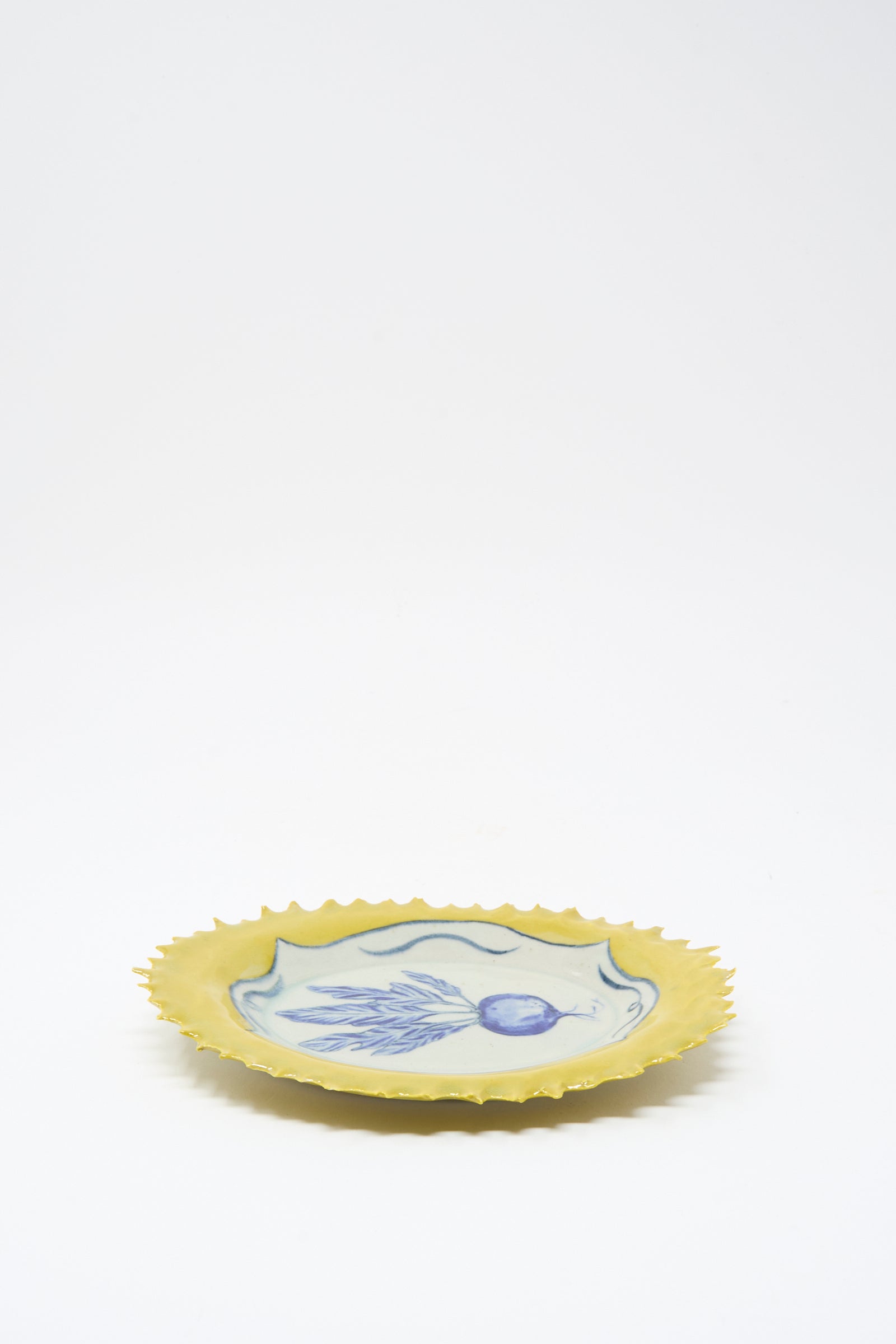 The Yellow Radish Plate by Ariel Clute is a handmade yellow ceramic plate with jagged edges and a blue and white fish illustration at the center, crafted in California, shown on a plain white background.