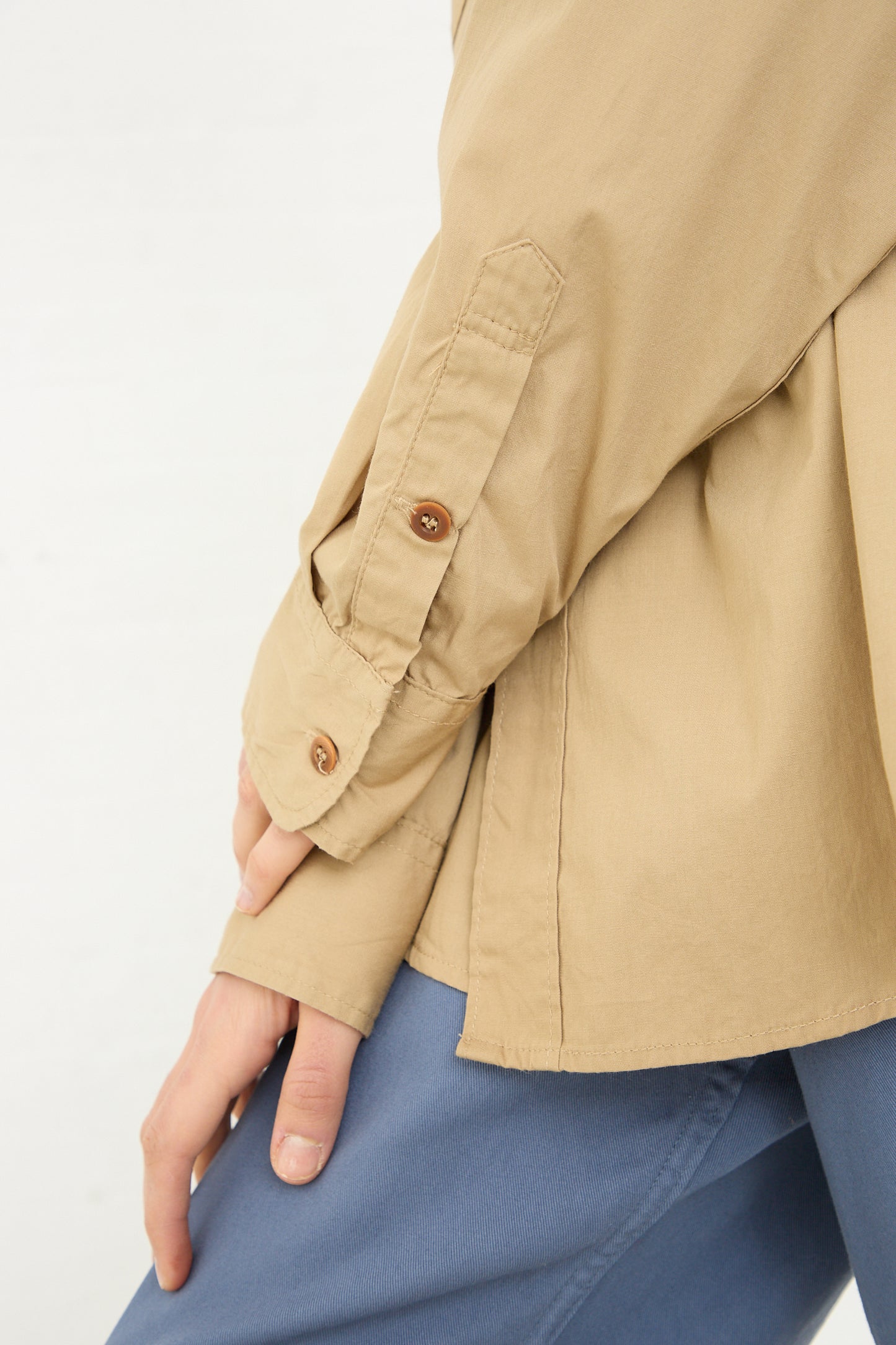 Close-up of a person wearing an As Ever 101 Shirt in 40's Khaki vintage jacket with a buttoned cuff, standing with their hand on their hip, wearing blue trousers.