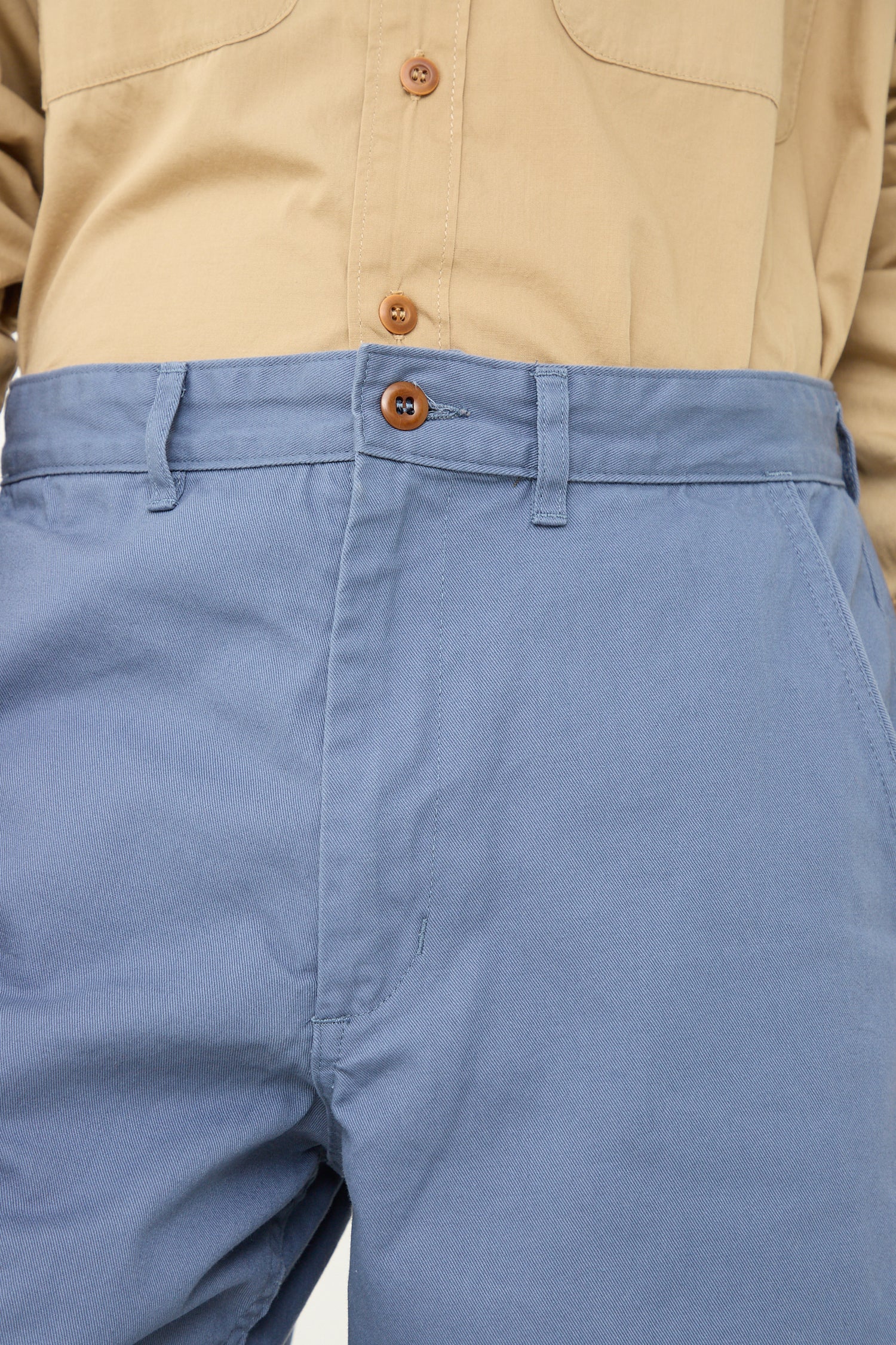 A close-up view of a person wearing beige and blue. The beige As Ever chino shirt is tucked into vintage military pants, which are secured by a button at the waistband.