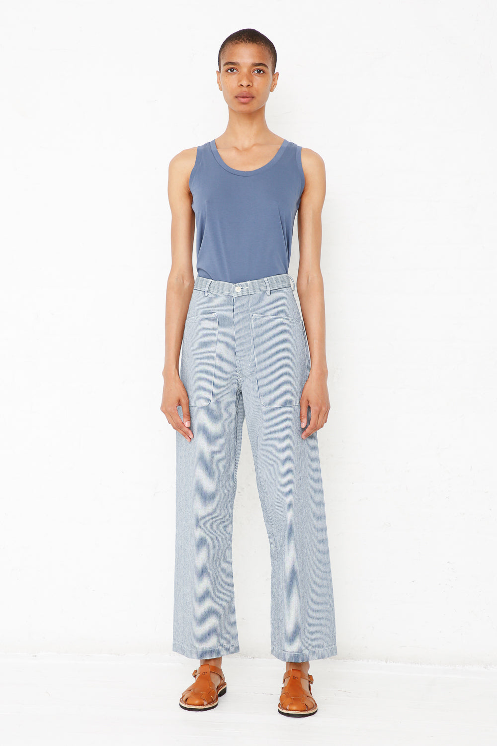 Wearing the Brancusi Pant in Engineer Stripe by As Ever, a person stands against a white background in a blue tank top and brown sandals with a neutral expression.