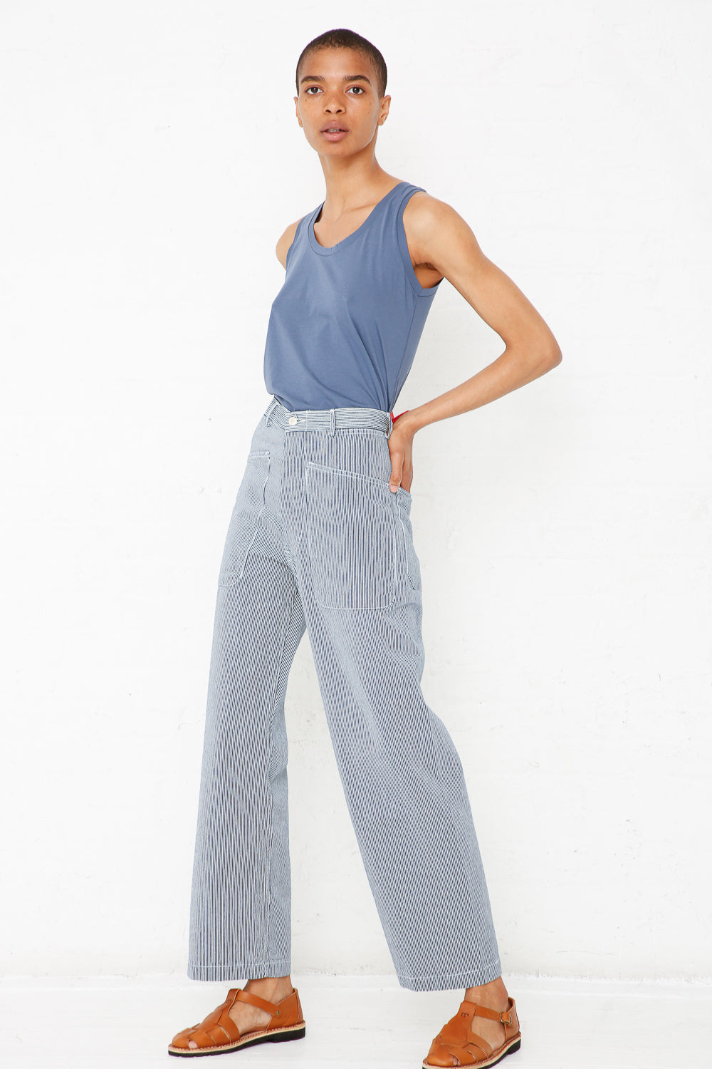 A person with short hair is standing sideways against a plain white background, wearing the Brancusi Pant in Engineer Stripe from As Ever, paired with a blue tank top.