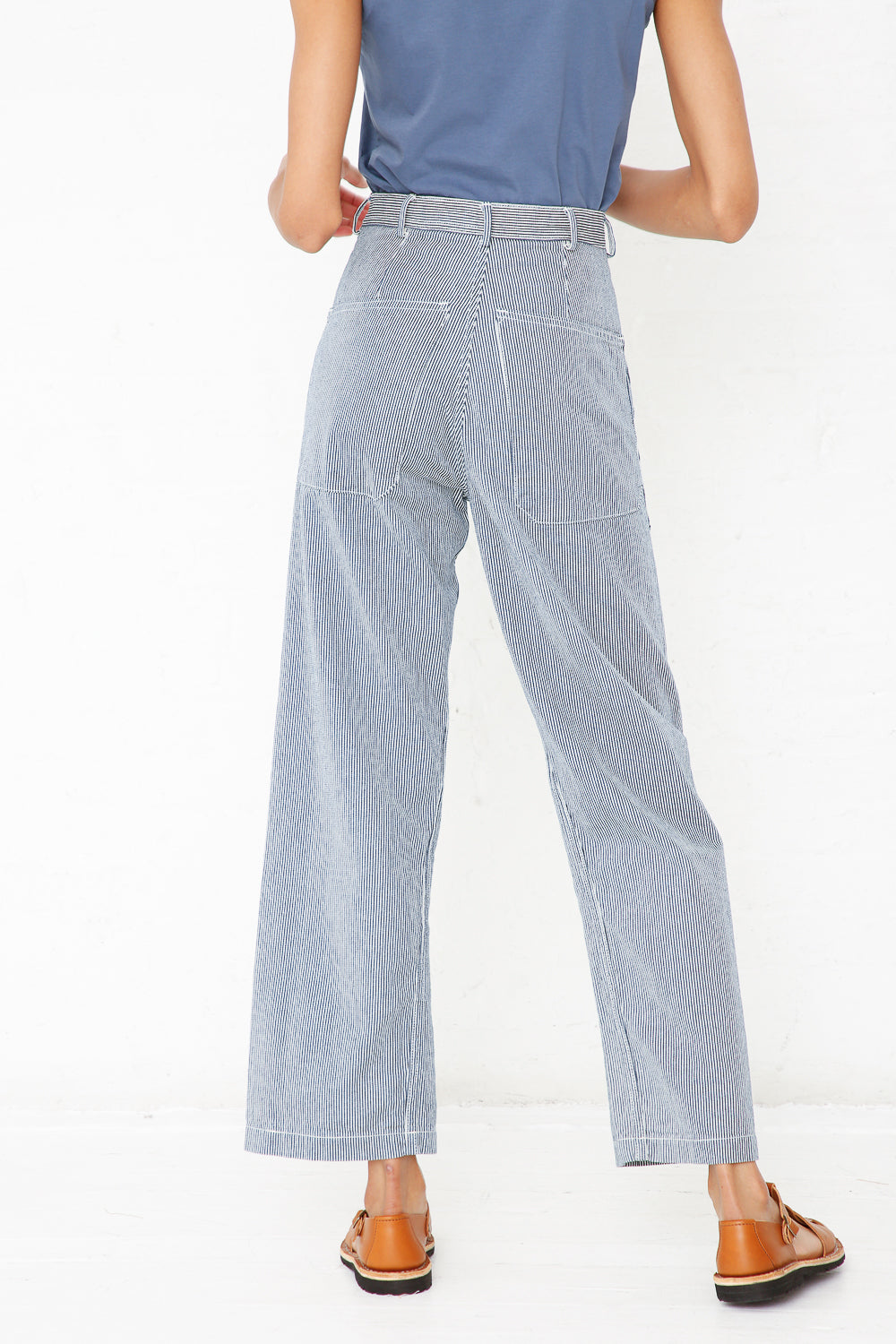 Wearing the As Ever Brancusi Pant in Engineer Stripe, a person stands with their back to the camera. The wide-leg silhouette is paired with brown shoes and a blue shirt, set against a plain white background.