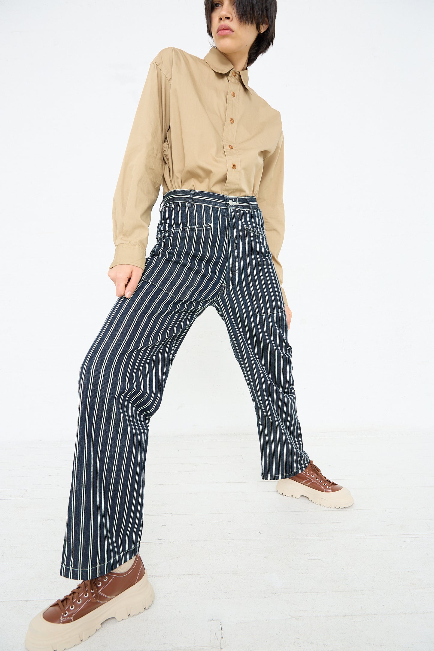 A person stands posing in a beige button-up shirt and high waisted navy blue pinstripe Brancusi Pant in Indigo Denim Stripe, paired with tan and brown platform shoes, against a white background. Brand Name: As Ever