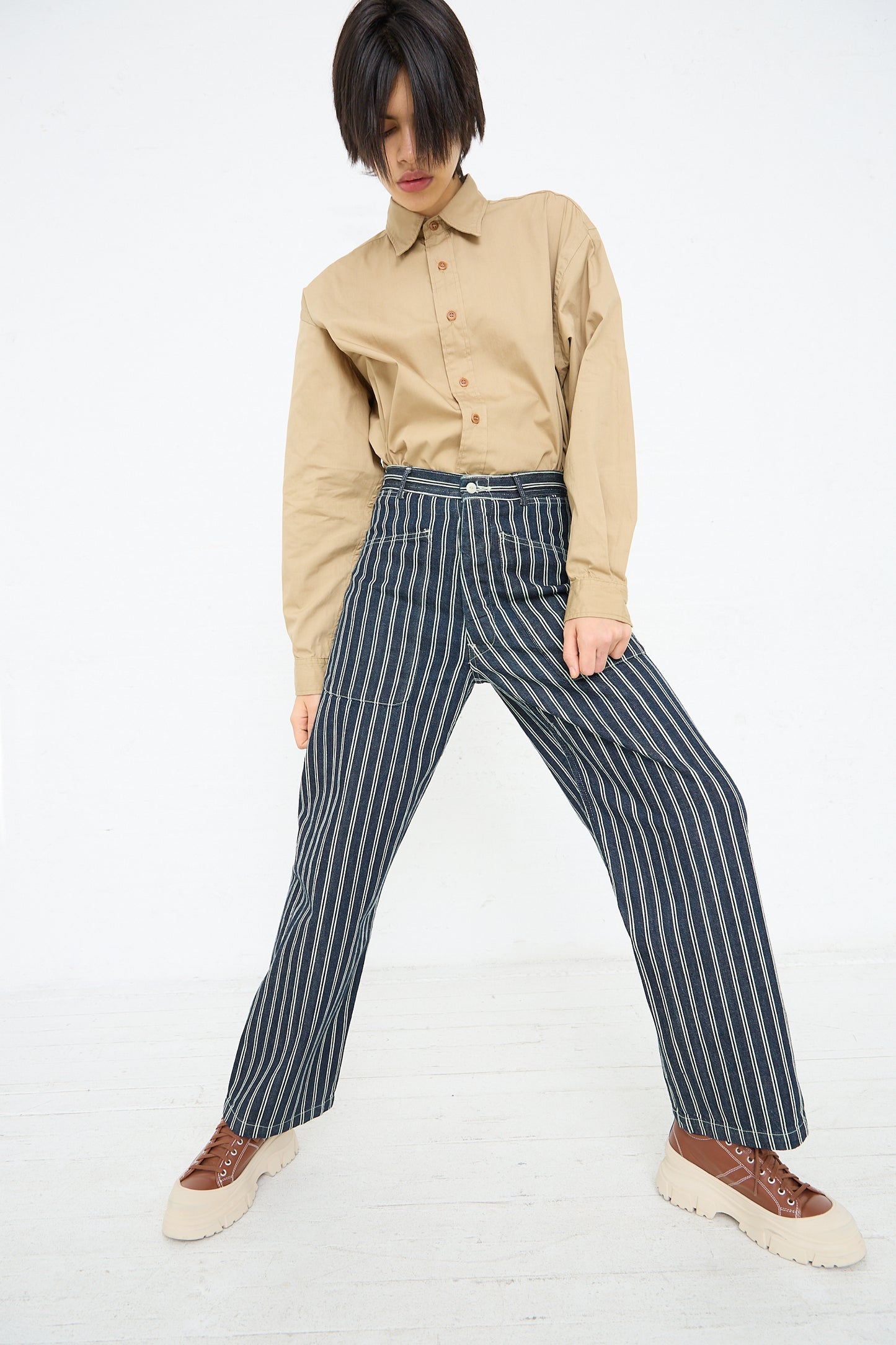 A person stands against a white background, wearing a beige button-up shirt, As Ever's Brancusi Pant in Indigo Denim Stripe, and two-toned shoes. They are facing forward with their hands.