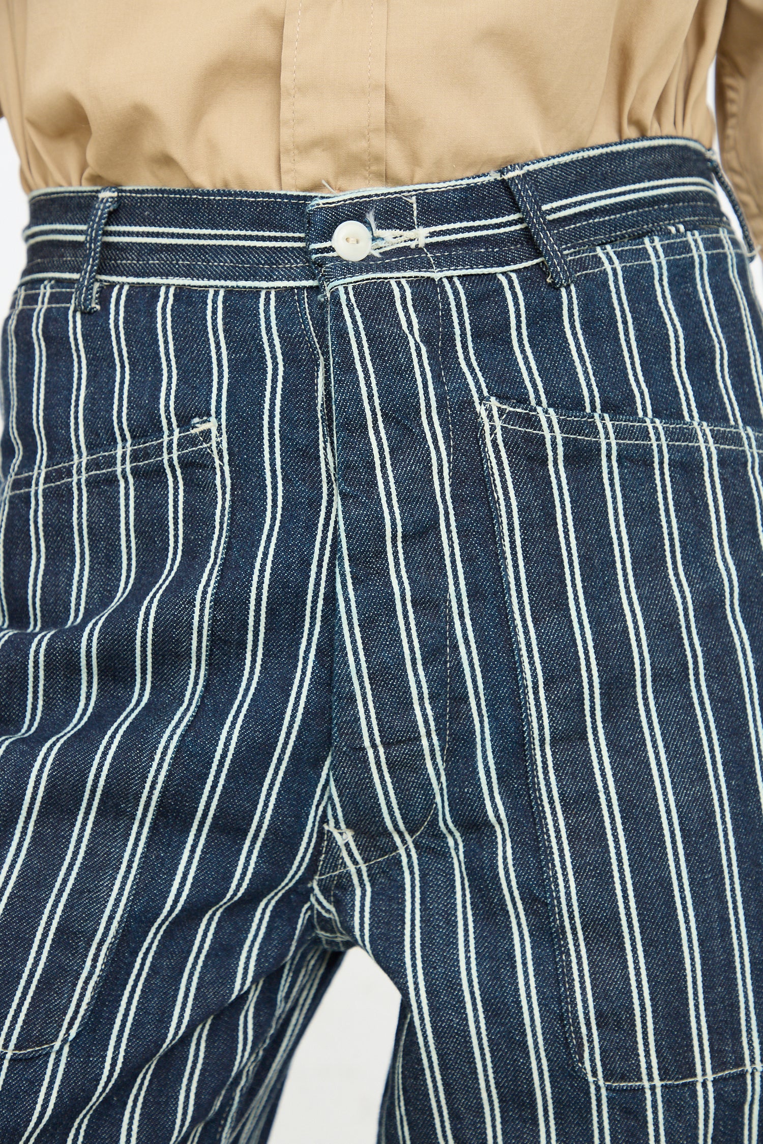 A close-up image of a person wearing As Ever's Brancusi Pant in Indigo Denim Stripe and a beige shirt tucked in at the waistline.