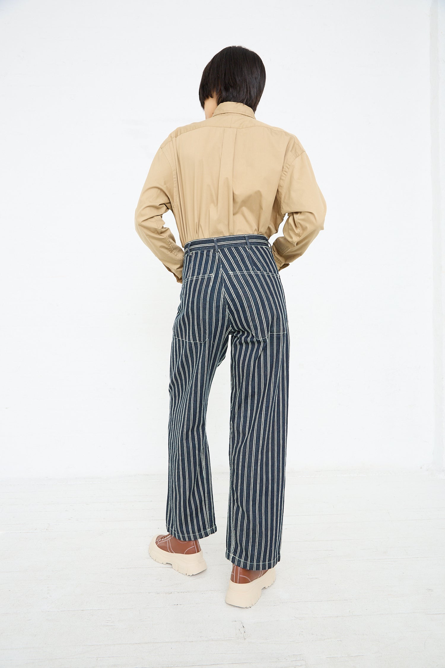 A person standing with their back to the camera, wearing a beige shirt and As Ever's Brancusi Pant in Indigo Denim Stripe trousers, paired with beige shoes, against a white background.