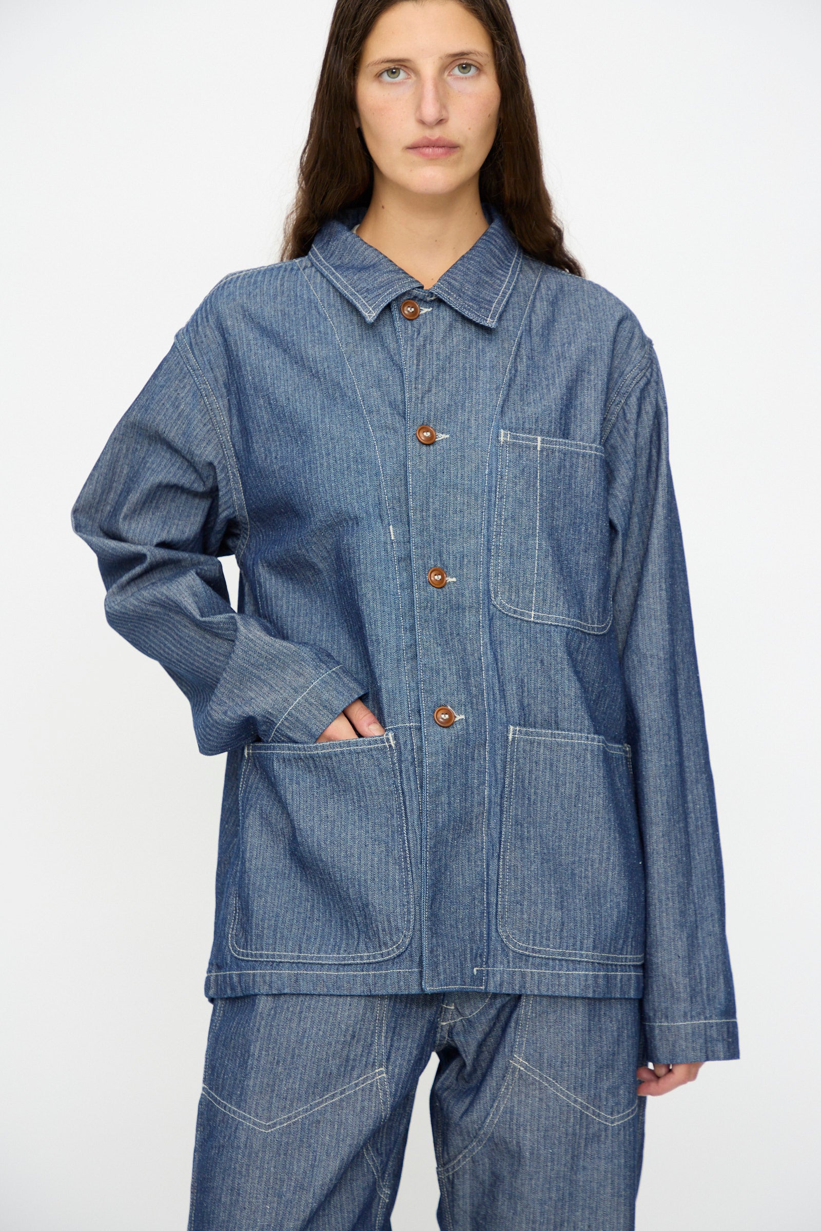 A person wears the As Ever Henri Jacket in HBT Denim— a loose-fit, unisex work jacket inspired by 1940s military styles, featuring front pockets and matching pants—standing against a plain background.