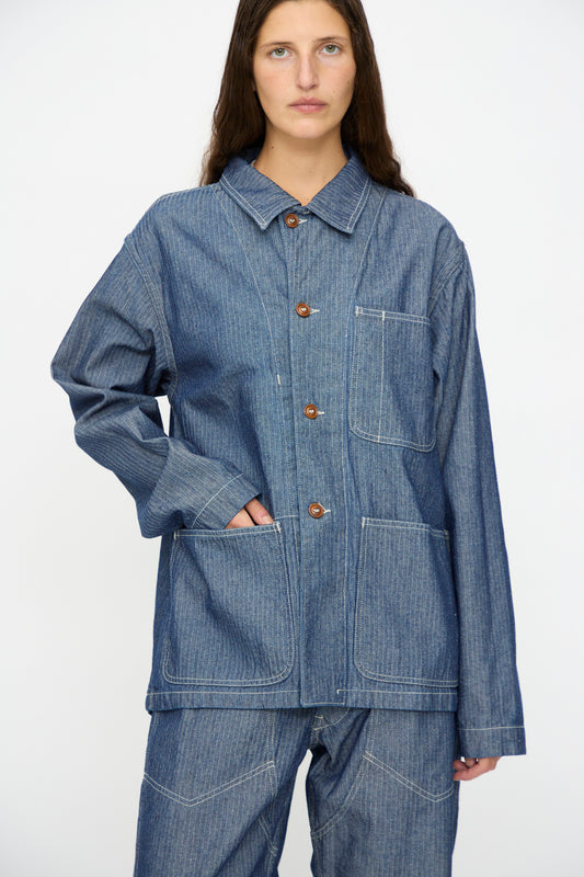A person wears the As Ever Henri Jacket in HBT Denim— a loose-fit, unisex work jacket inspired by 1940s military styles, featuring front pockets and matching pants—standing against a plain background.