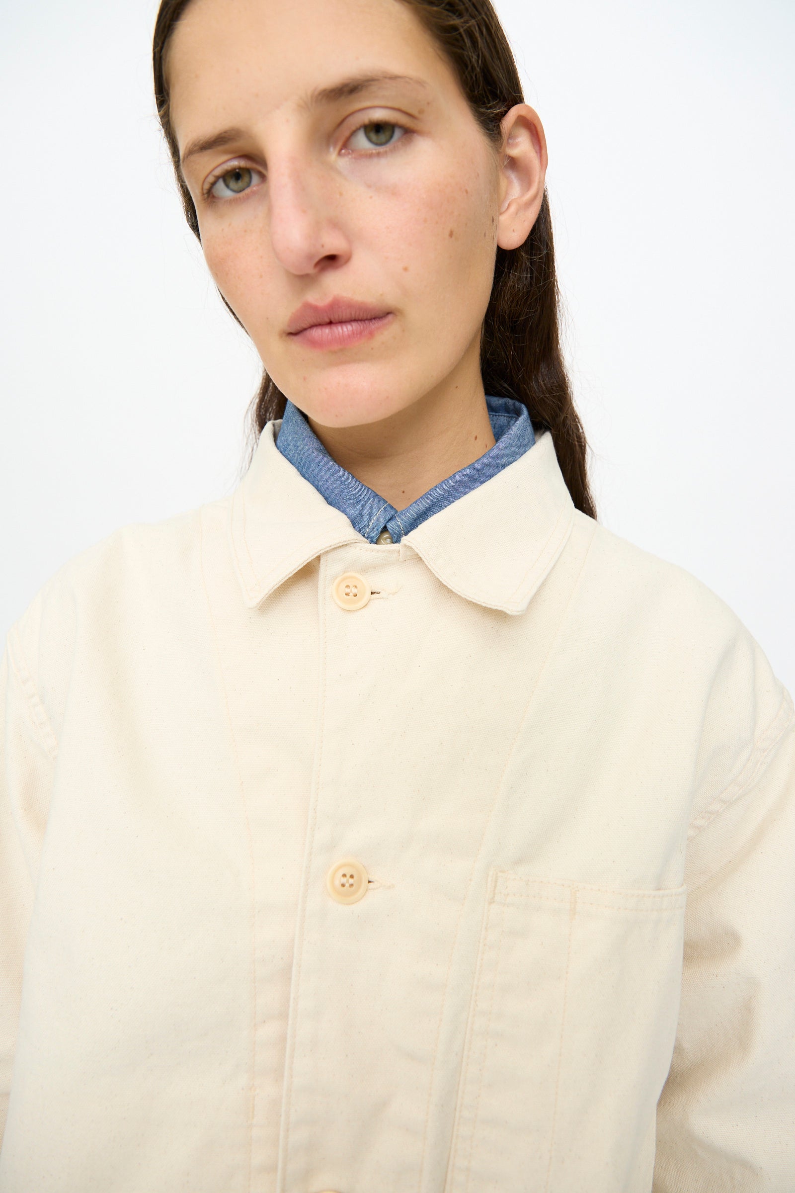 A person with long brown hair wears the As Ever Henri Jacket in Natural over a blue shirt, standing against a plain white background.