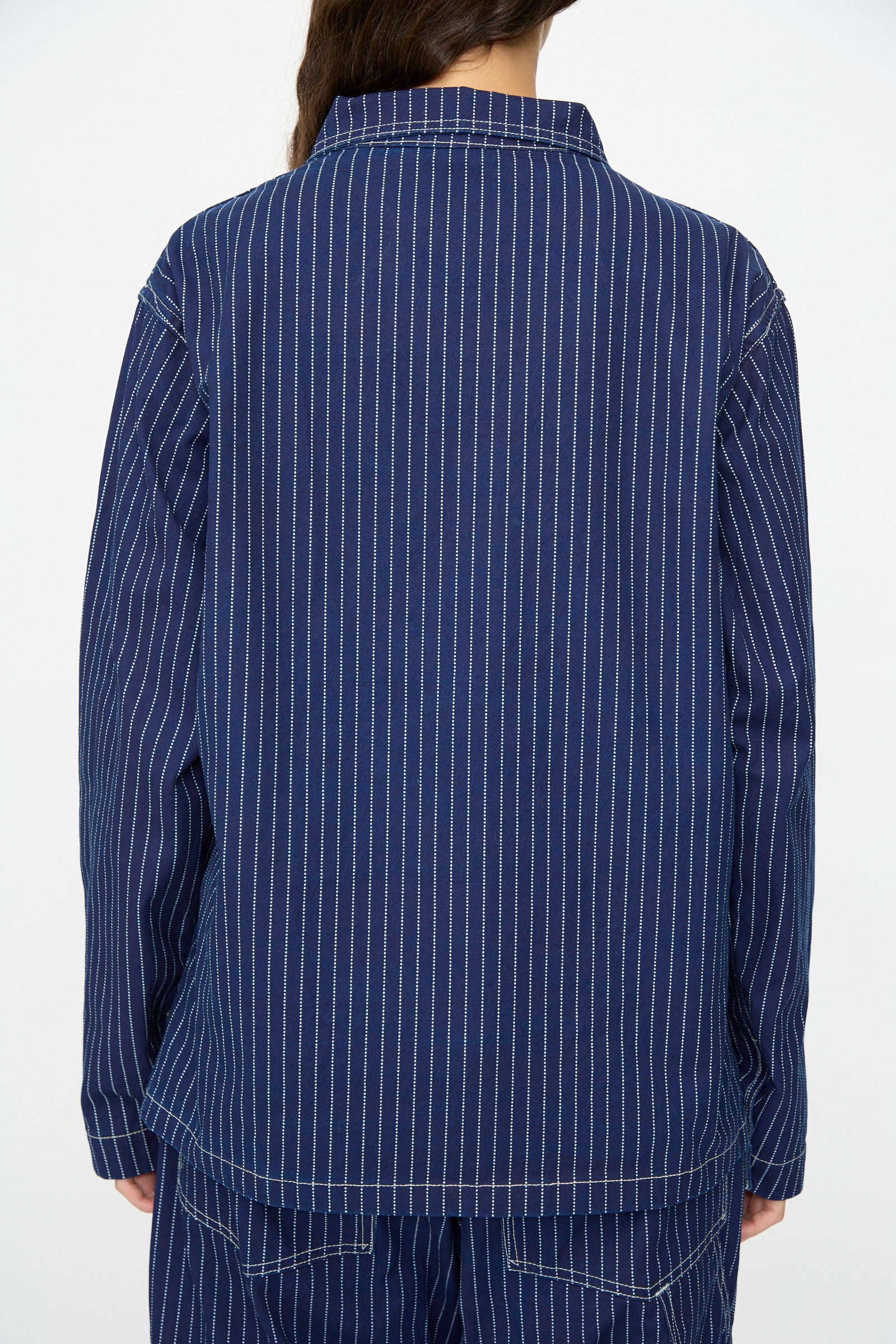 A person is seen from the back wearing the As Ever Henri Jacket in Wabash, featuring indigo fabric with white vertical pinstripes, paired with matching blue pants for a utilitarian look.