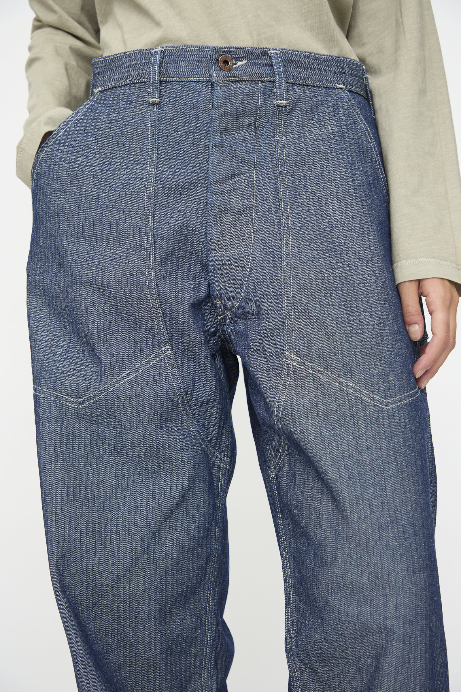 A person wears a light long-sleeve shirt with the Henri Pant in HBT Denim by As Ever—high-waisted, vintage military pants made from blue denim with visible white stitching.