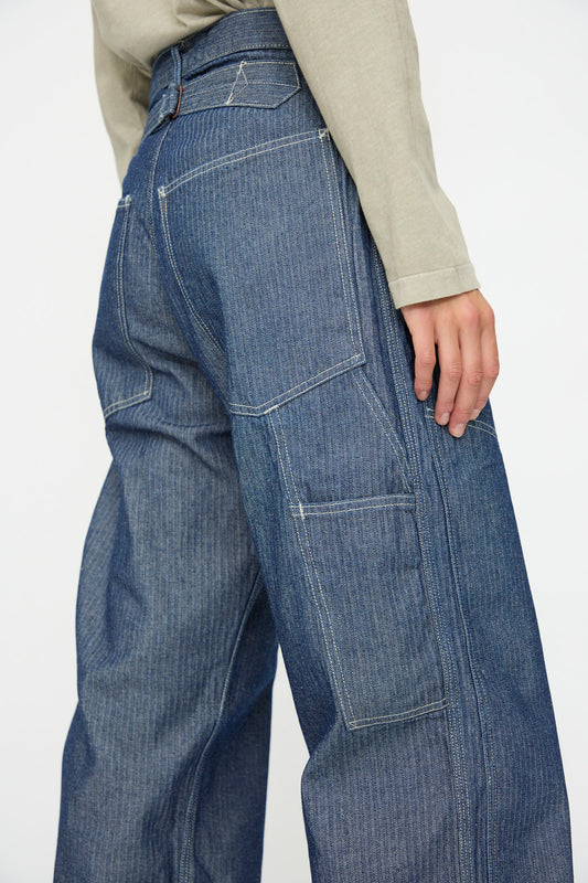 Model wears the As Ever Henri Pant in HBT Denim, featuring oversized blue denim with large pockets, paired with a light-colored long-sleeve shirt; shown waist down from a side view.