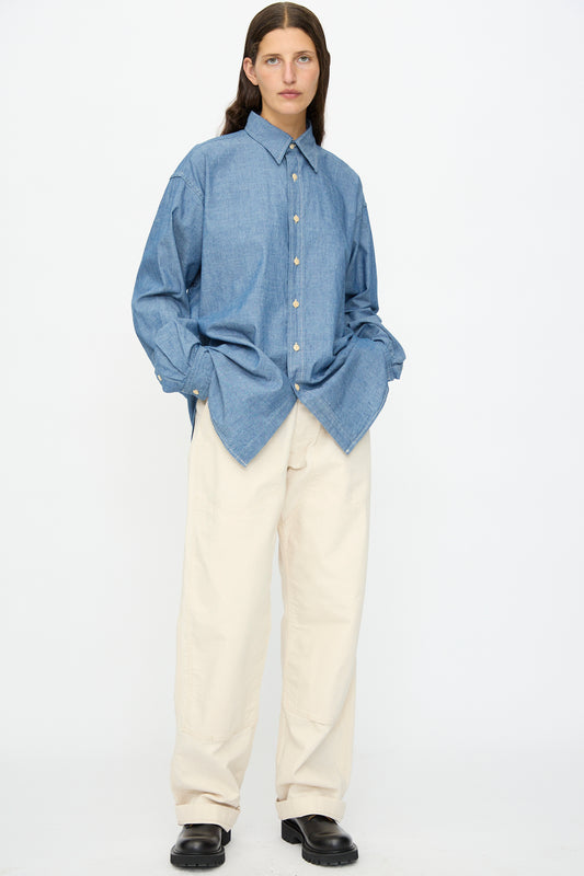 A person wearing a loose blue button-up shirt, As Ever Henri Pant in Natural, and black shoes stands against a plain white background.