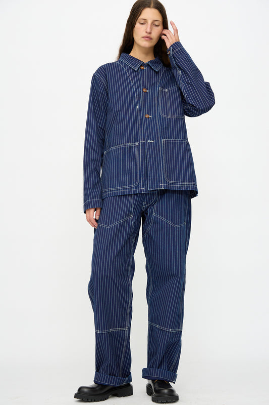 A person stands against a white background, wearing the Henri Pant in Wabash by As Ever with a matching blue pinstripe jacket and black shoes, touching their hair with one hand.