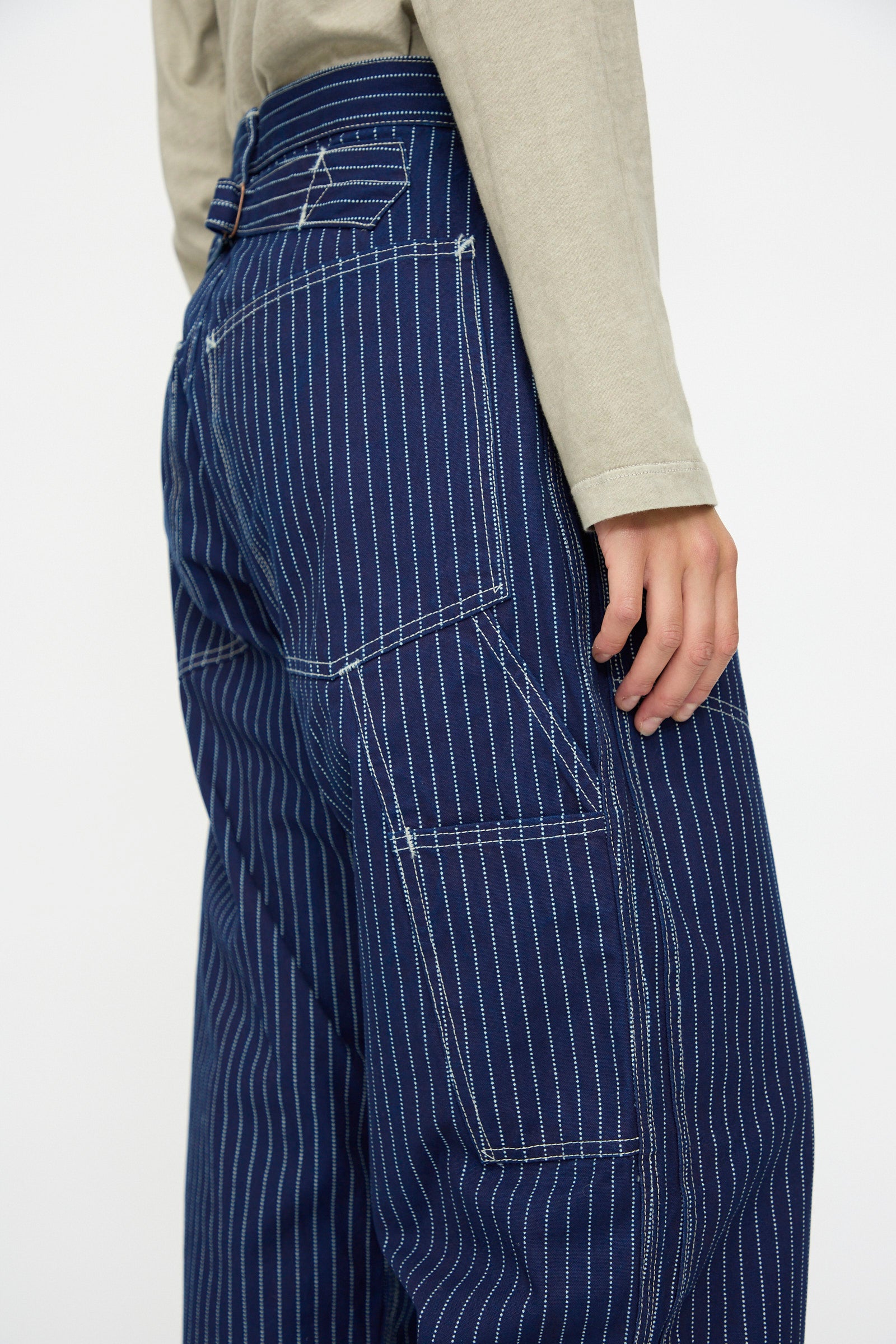A person shown from the waist down at a side angle wears As Ever's Henri Pant in Wabash, which features loose-fitting, blue pinstriped vintage military styling with visible stitching, paired with a beige long-sleeve shirt.