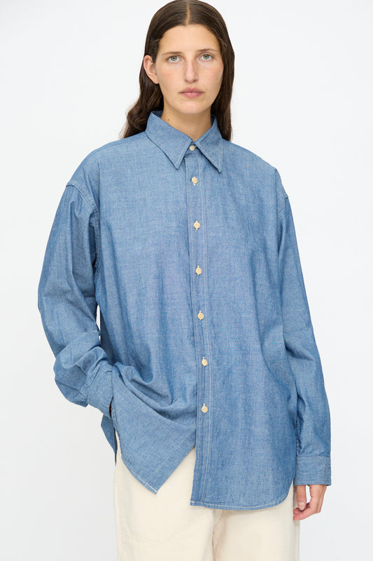 A person wears the As Ever Largo Shirt in Chambray, paired with off-white pants, standing against a plain white background.