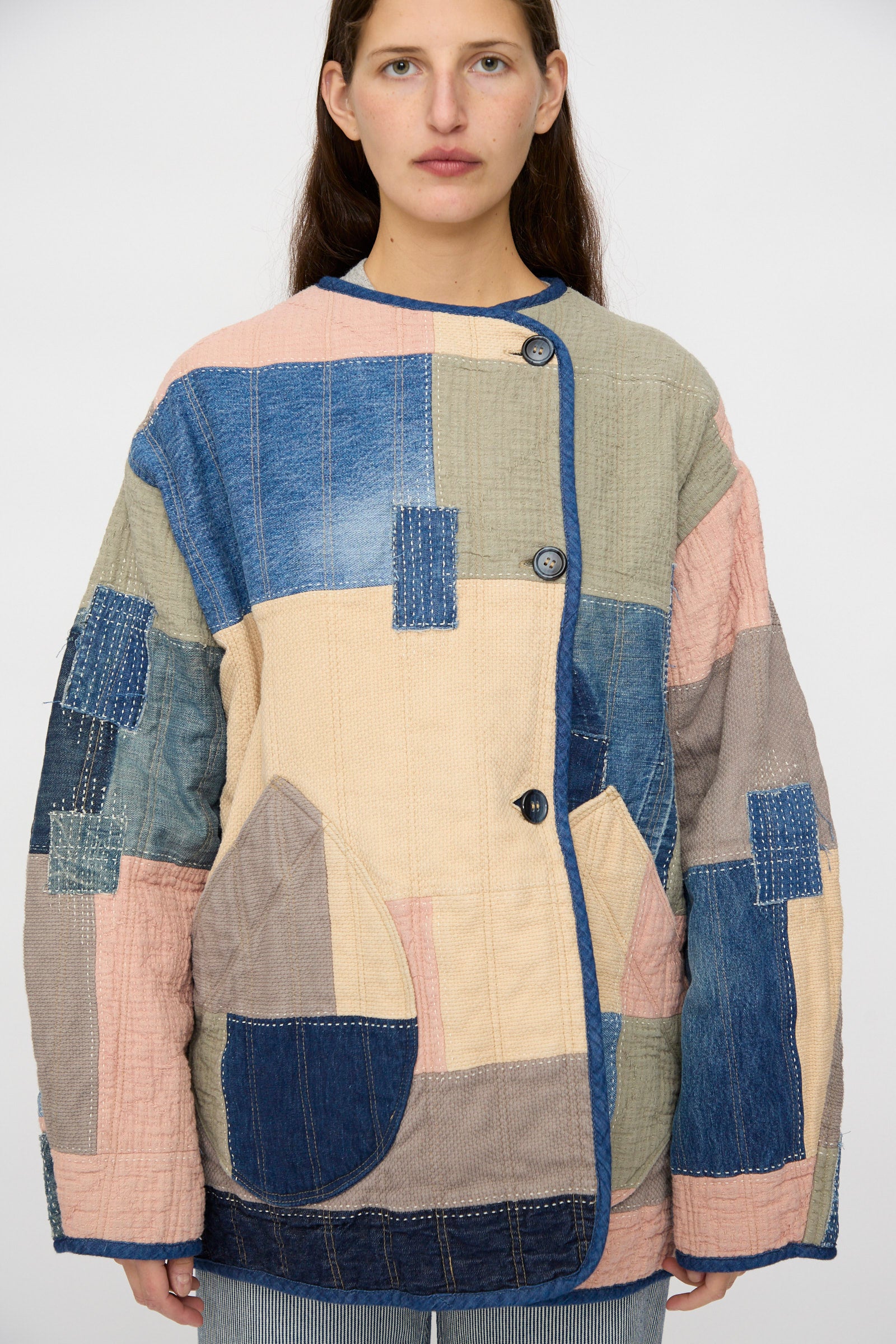 A person wears the As Ever Quilt Coat in Color Block Boro, a loose jacket made from vintage denim and Japanese cotton with pastel patchwork squares, standing against a plain background.