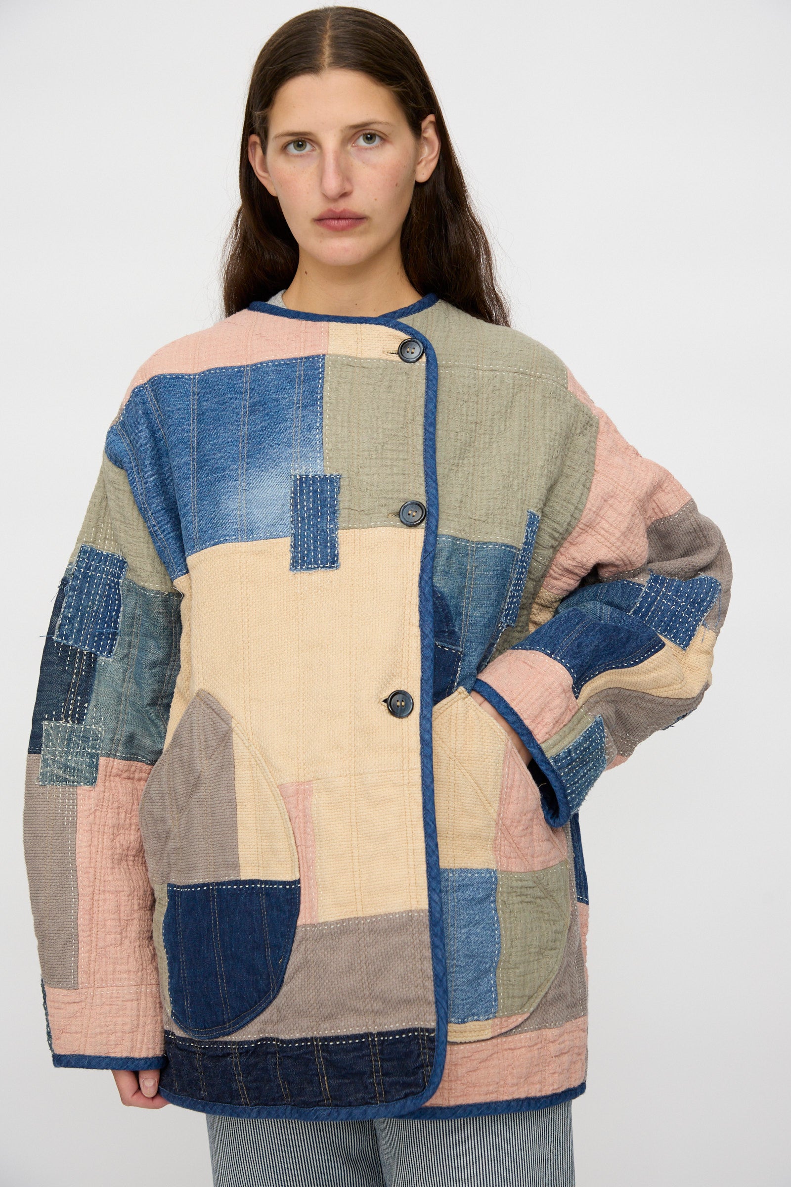 A woman stands against a plain background wearing As Ever's Quilt Coat in Color Block Boro, featuring blue, beige, pink, and green Japanese cotton squares, paired with striped pants.