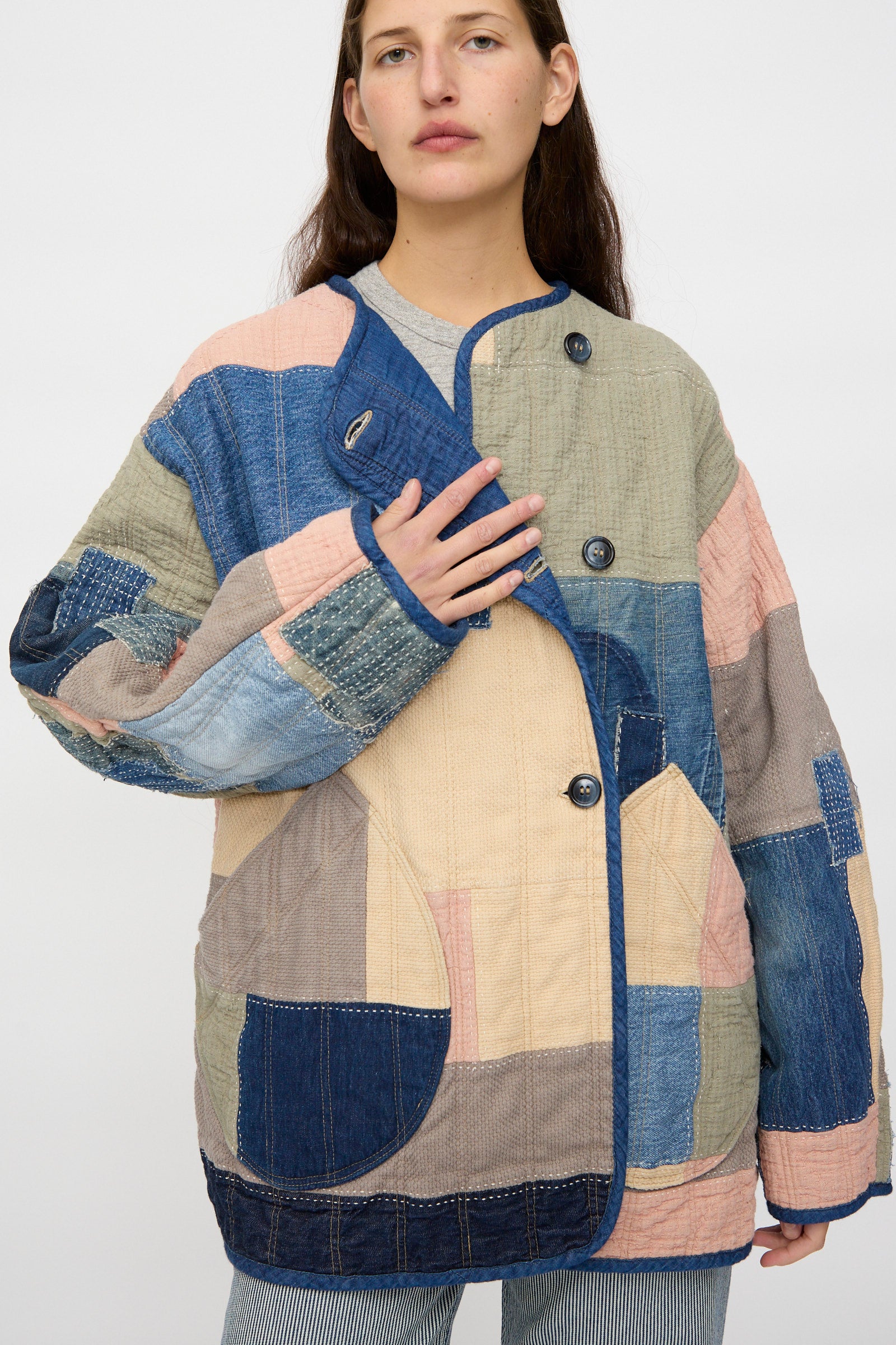 Someone wears the As Ever Quilt Coat in Color Block Boro, a patchwork jacket made from Japanese cotton in blue, beige, pink, and gray hues, standing against a plain background.