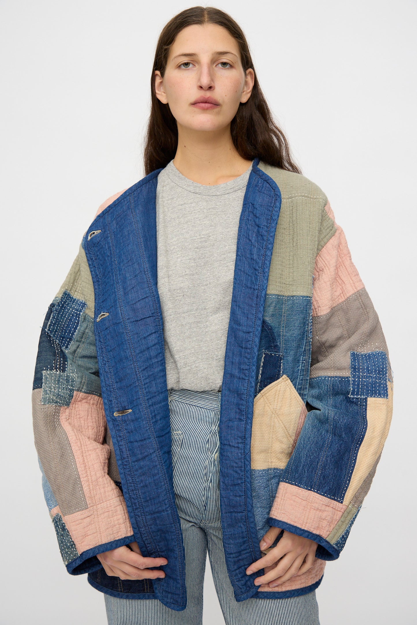 A person faces forward against a plain background, wearing the As Ever Quilt Coat in Color Block Boro— a vintage-style denim jacket with patchwork in blue, pink, and beige shades—over a grey t-shirt and striped pants.