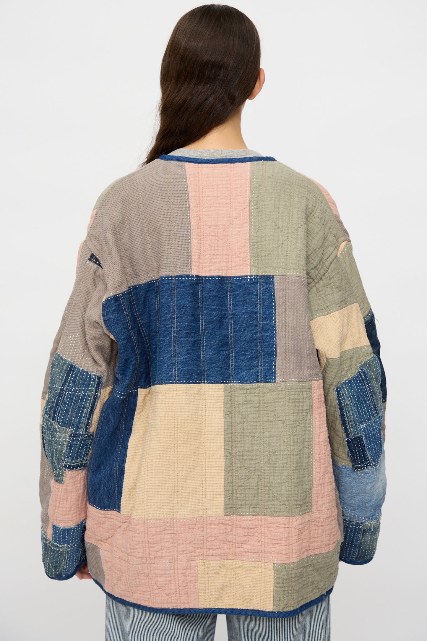 A person with long brown hair, seen from behind, wears the As Ever Quilt Coat in Color Block Boro—crafted from Japanese cotton in blue, pink, beige, and gray—paired with light blue jeans.