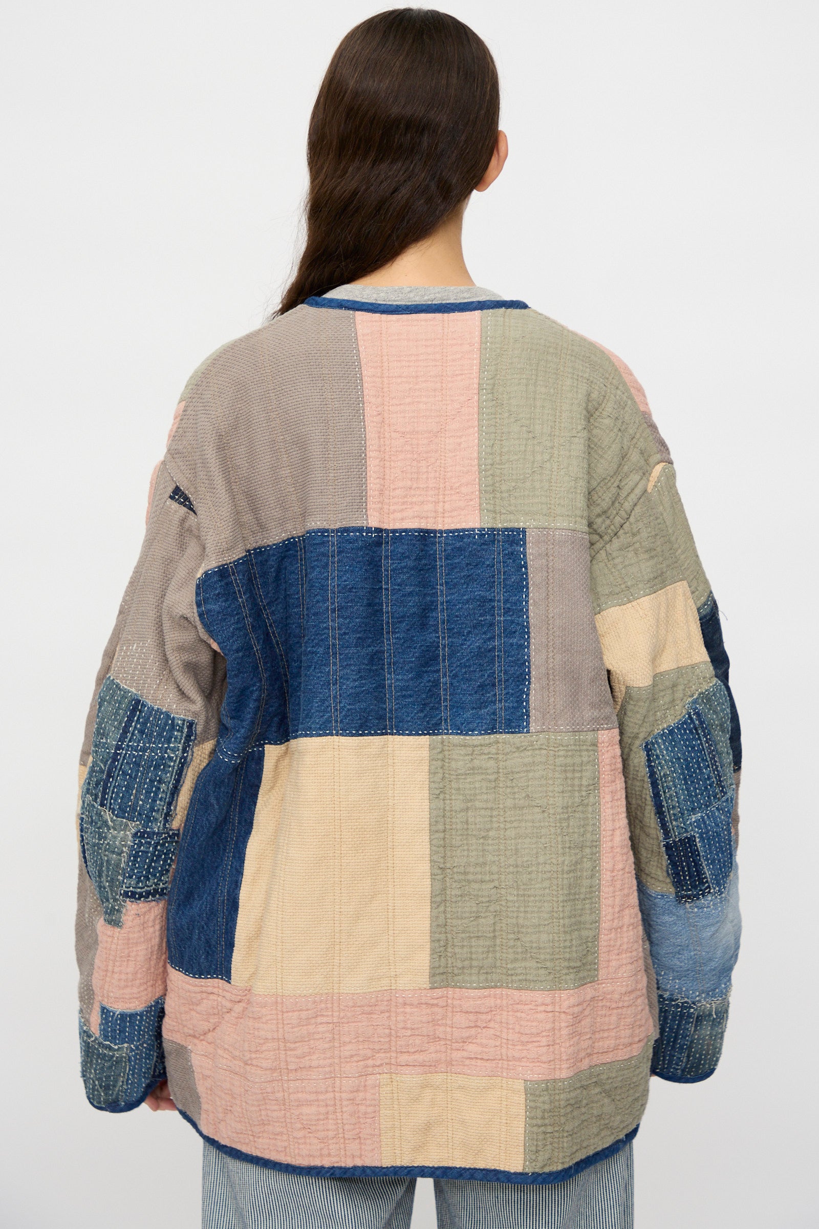 A person with long brown hair, seen from behind, wears the As Ever Quilt Coat in Color Block Boro—crafted from Japanese cotton in blue, pink, beige, and gray—paired with light blue jeans.