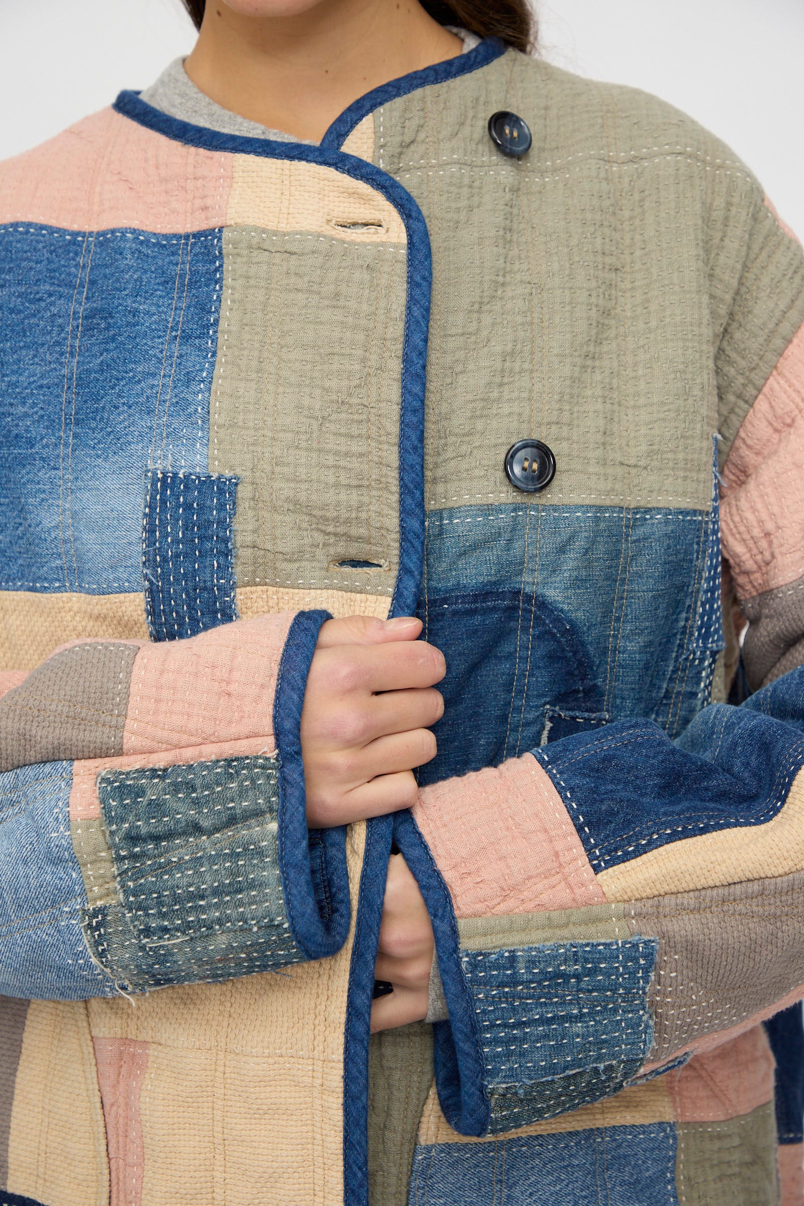 A person wearing the As Ever Quilt Coat in Color Block Boro, featuring patchwork quilting, visible stitching, and blue buttons, is shown holding the front of the jacket closed; only their torso and part of their face are visible.