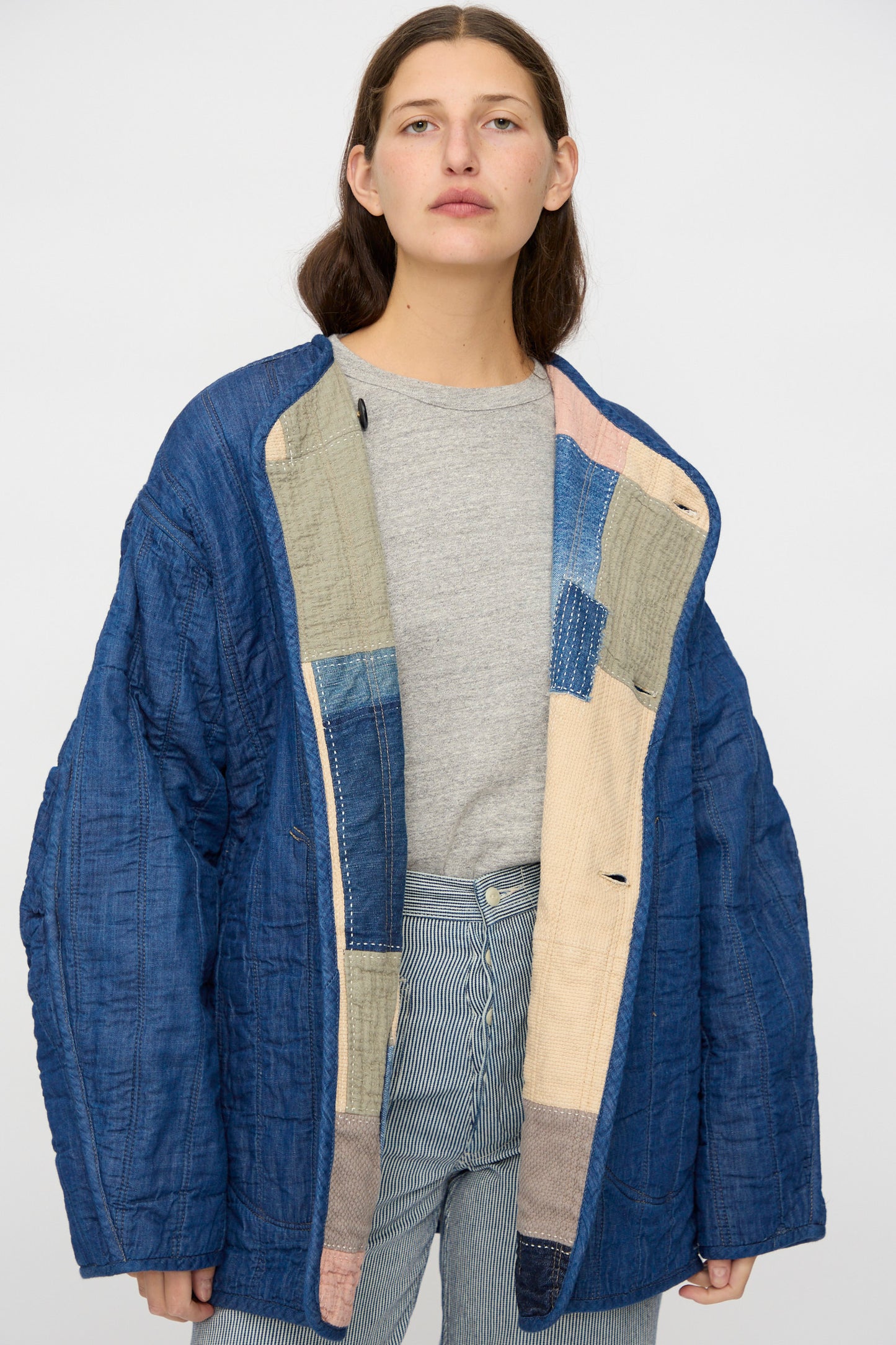 A person wears the Quilt Coat in Color Block Boro by As Ever— a loose blue quilted jacket made from Japanese cotton— over a grey t-shirt and striped pants, standing against a plain background.
