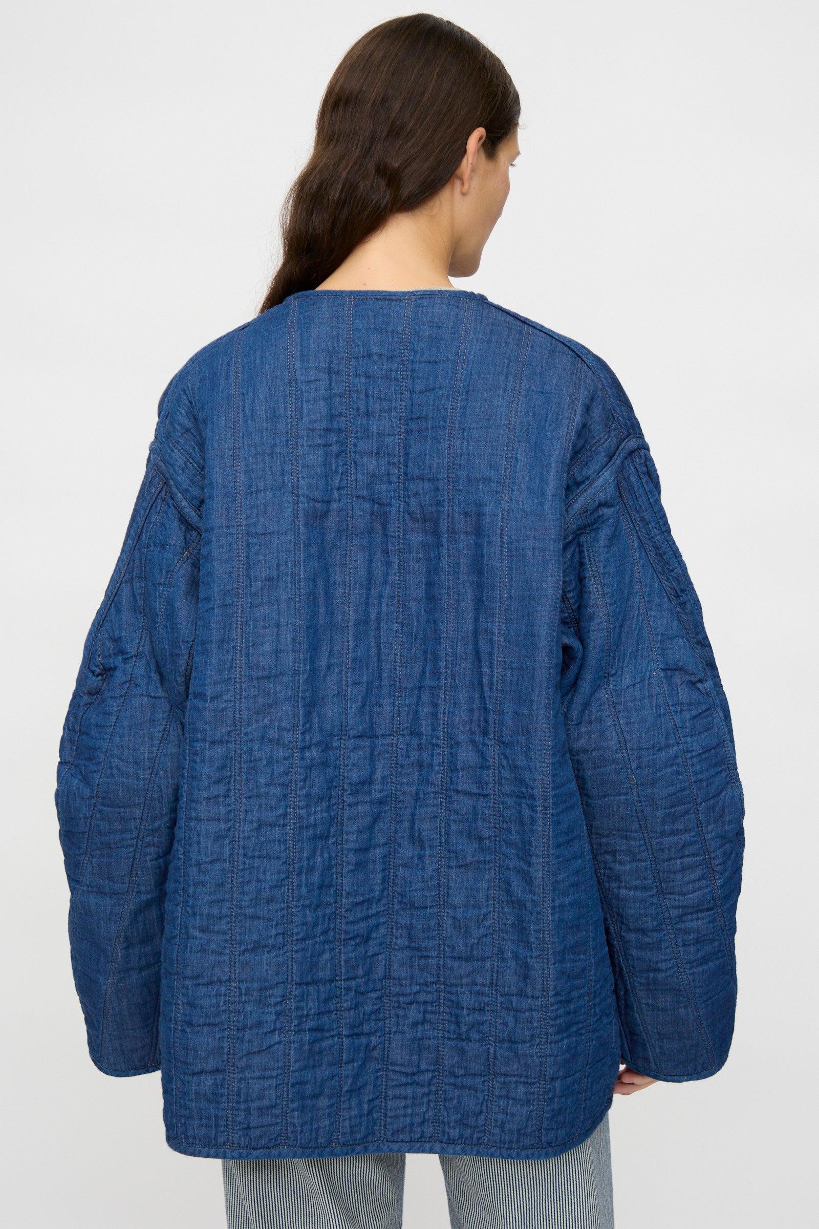 A person with long brown hair is seen from the back wearing the As Ever Quilt Coat in Color Block Boro, a loose Japanese cotton jacket, paired with light blue pinstriped pants against a plain background.