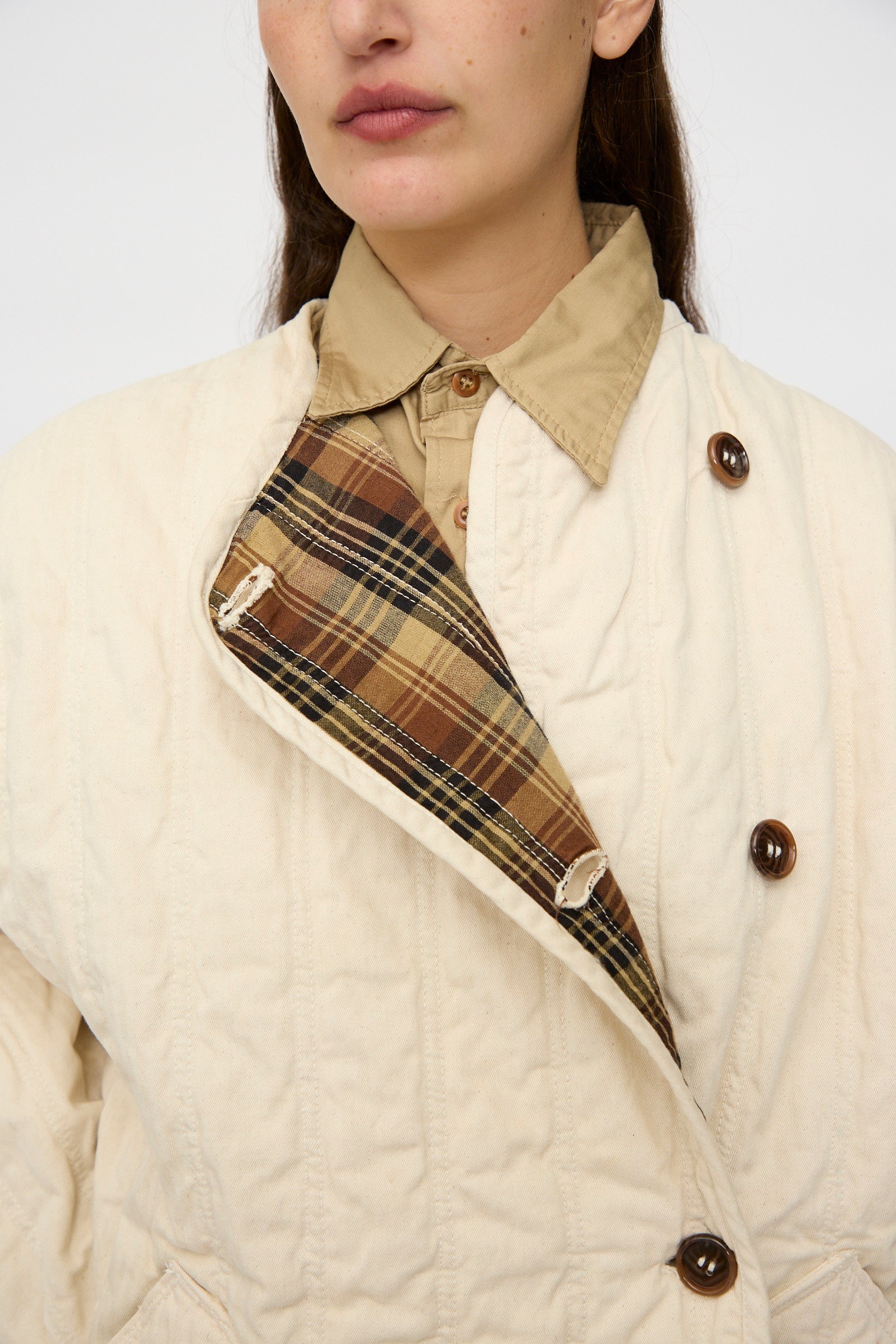 A person wears the As Ever Quilt Coat in HBT Natural—an oversized cream jacket with brown buttons, brown plaid lining, and soft cotton batting for warmth—over a beige collared shirt.