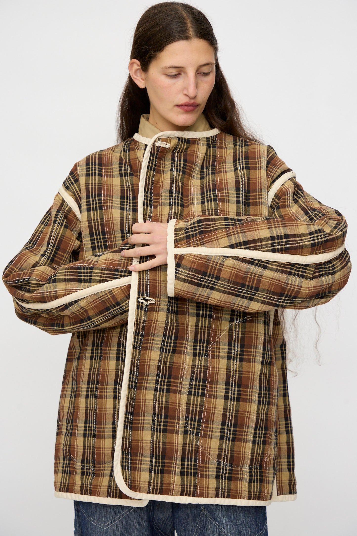 A person adjusts the front of the As Ever Quilt Coat in HBT Natural—an oversized fit brown plaid jacket with cotton batting and cream trim—while looking down against a plain background.